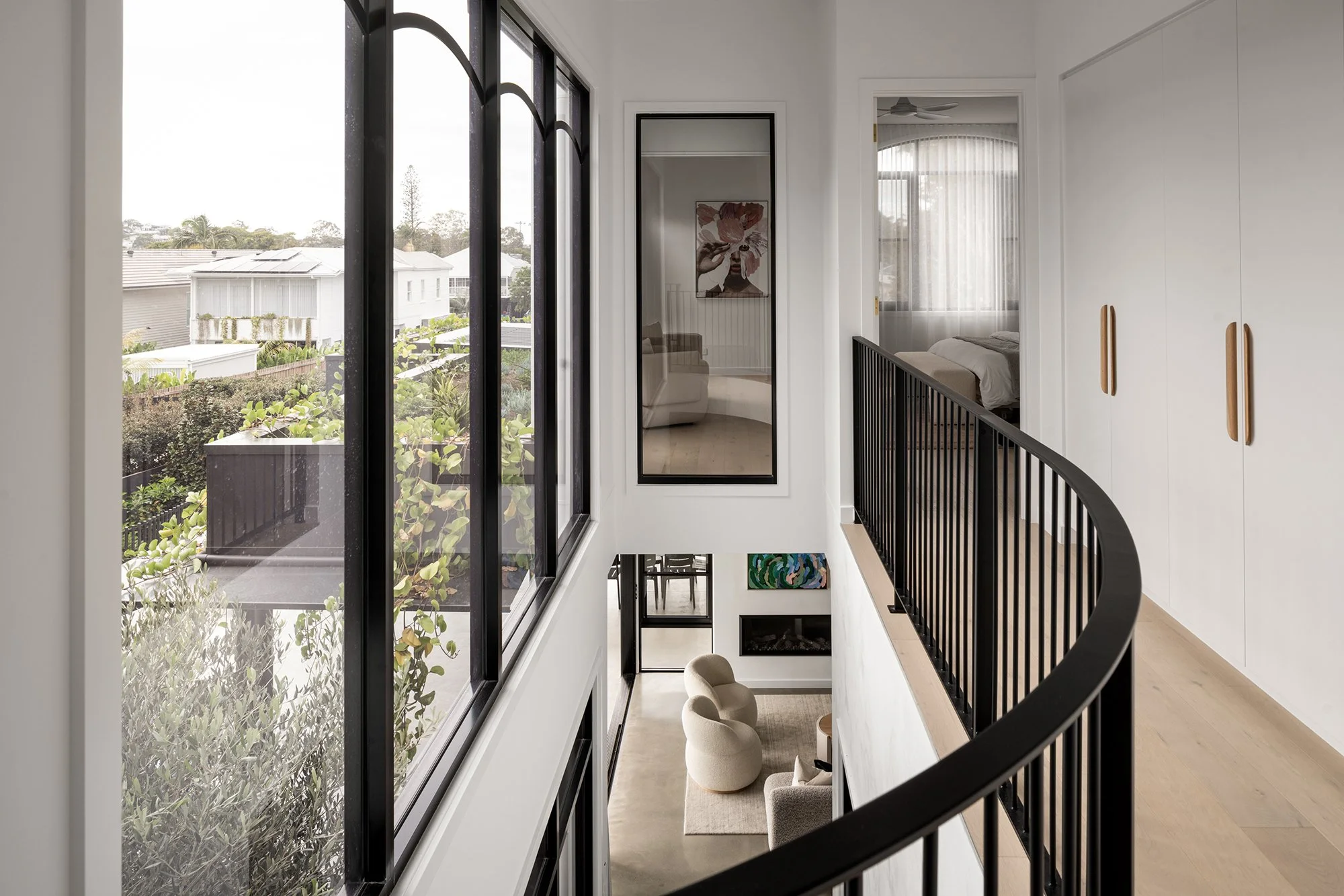 Interior view of a modern home with a balcony, large windows, a black railing, a sitting area with beige chairs, and a fireplace.