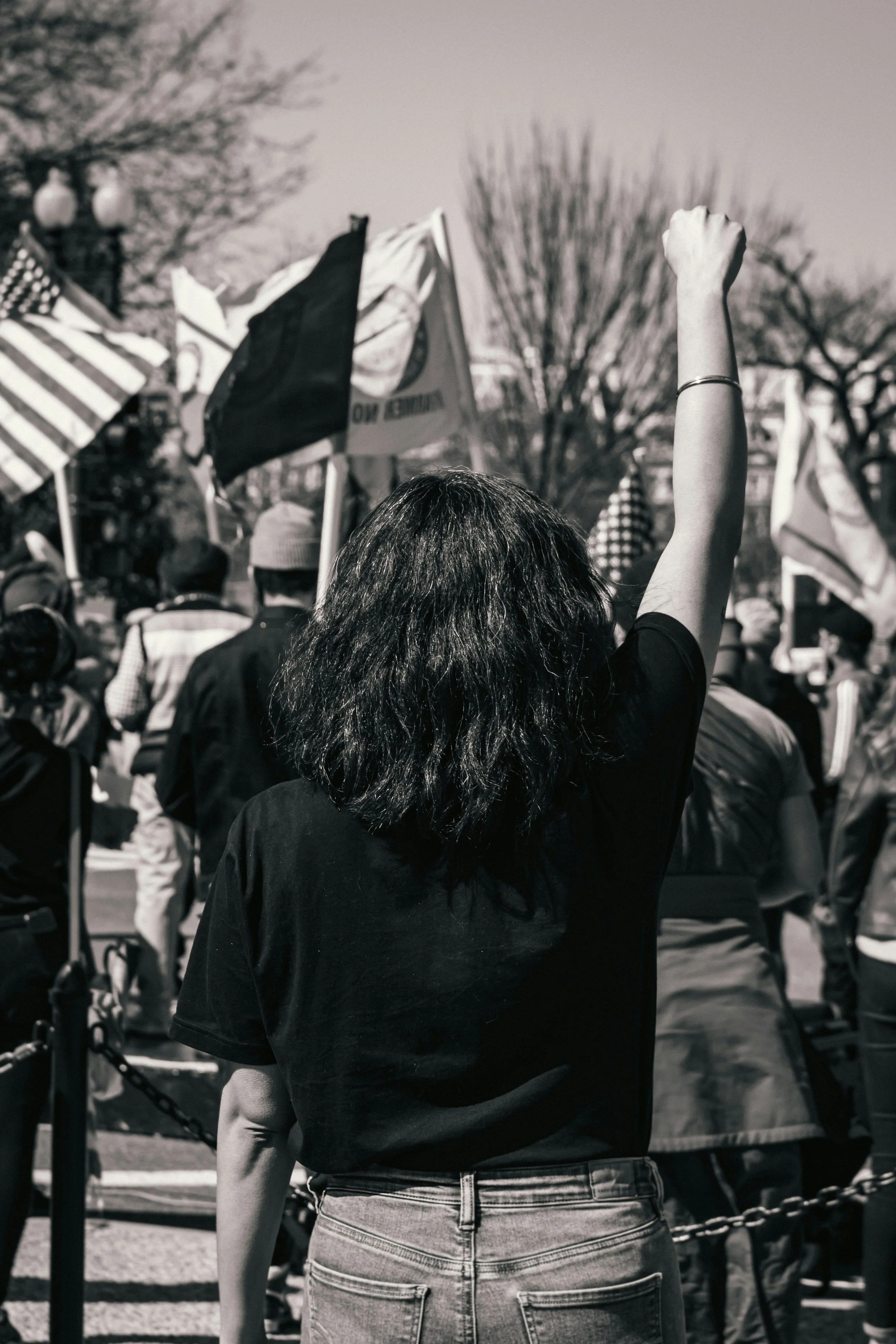 Une femme avec des cheveux bouclés, portant un t-shirt noir et un jean, le bras droit levé en l'air lors d'une manifestation avec des drapeaux en arrière-plan.