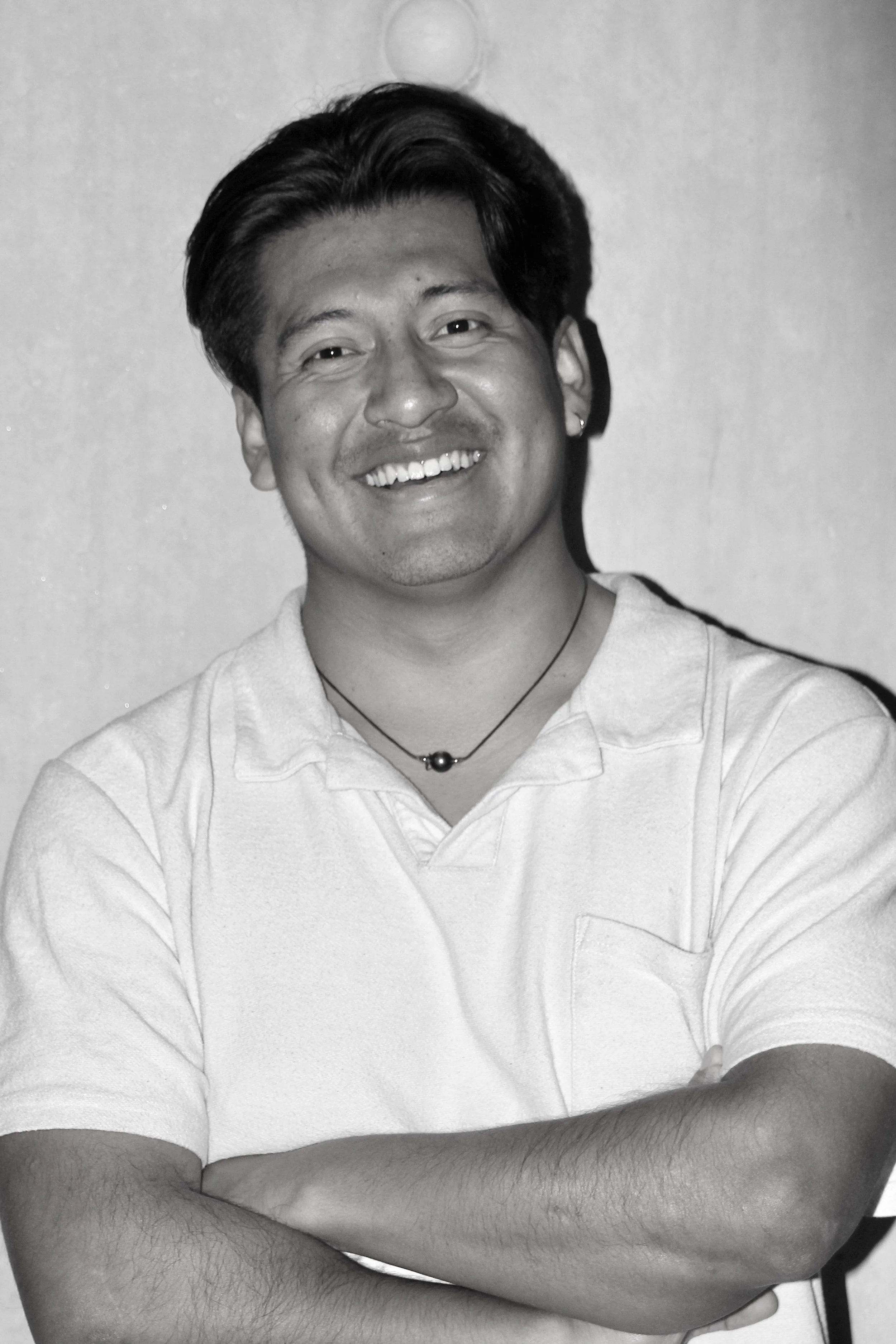 Un homme souriant avec les bras croisés portant un t-shirt blanc, portant un collier noir, en noir et blanc.