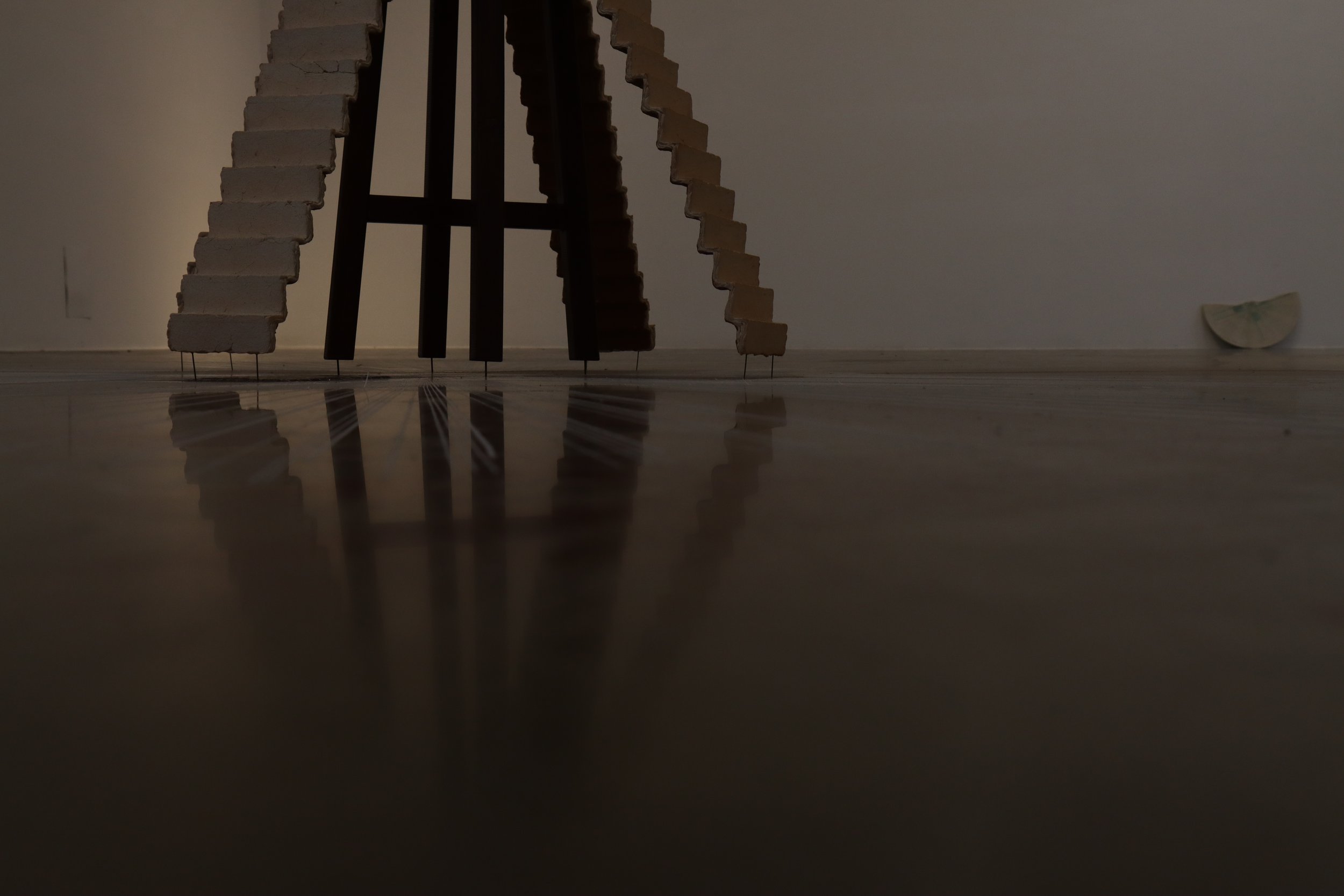 Modern art sculpture made of vertical wooden and metal beams, with a small white abstract object on the floor to the right, all reflected on a shiny wooden floor.