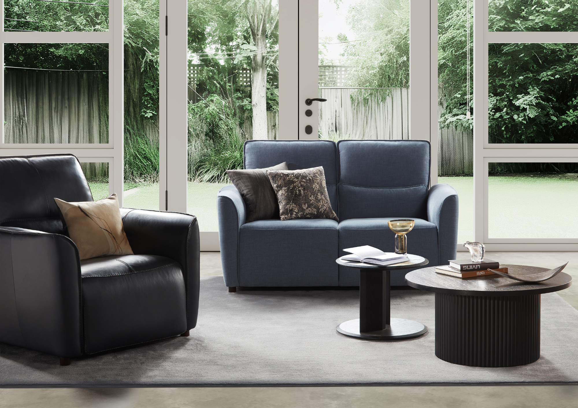 Living room with black leather armchair and blue couch, both with cushions, in front of large glass doors showing a lush green garden outside. Coffee tables with books, a glass, and decorative items are in the room.