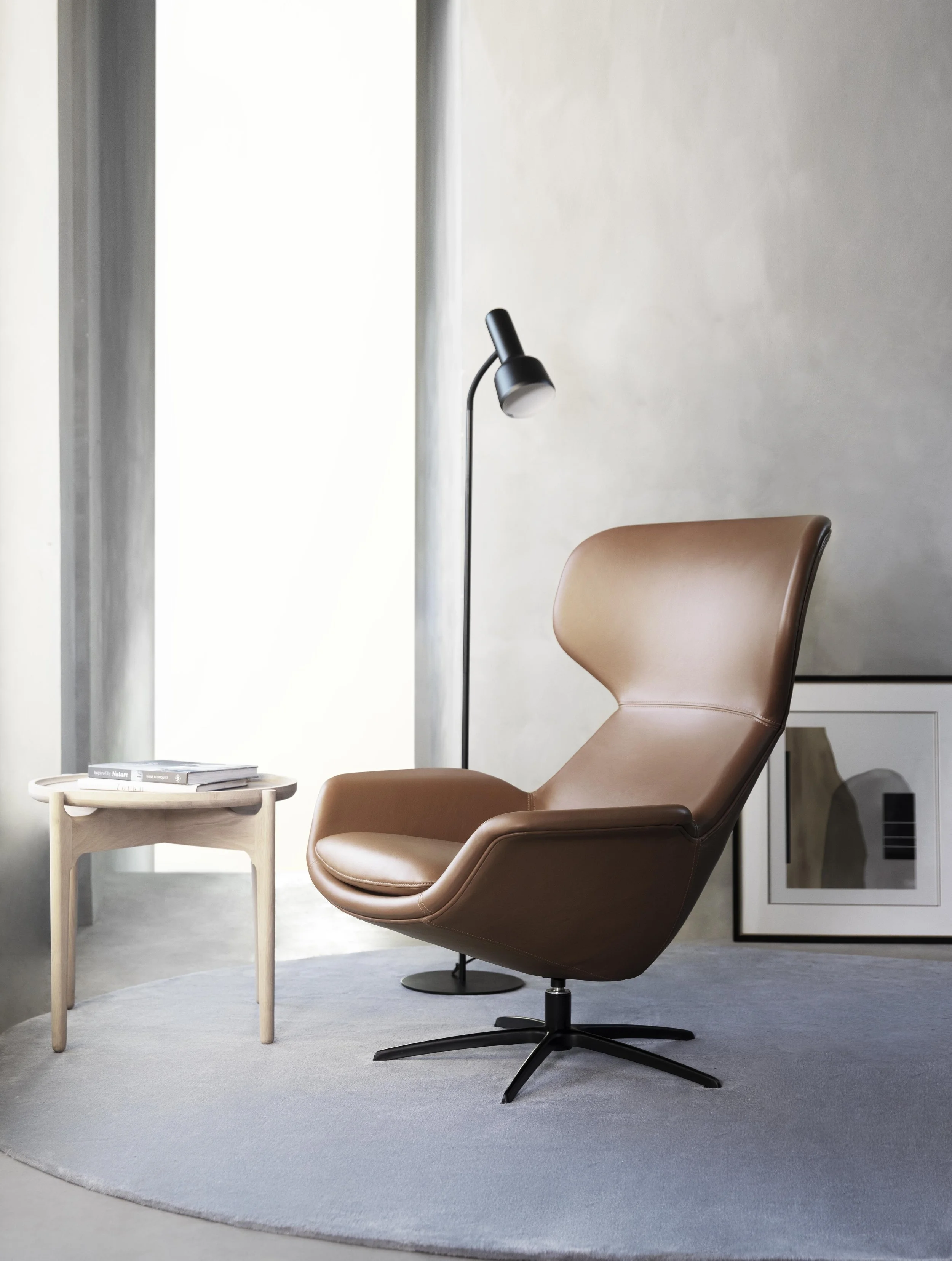 A cozy modern workspace featuring a brown leather swivel chair, a black floor lamp, a small white side table with books, a light gray rug, and a wall with framed artwork, illuminated by natural light from a nearby window.