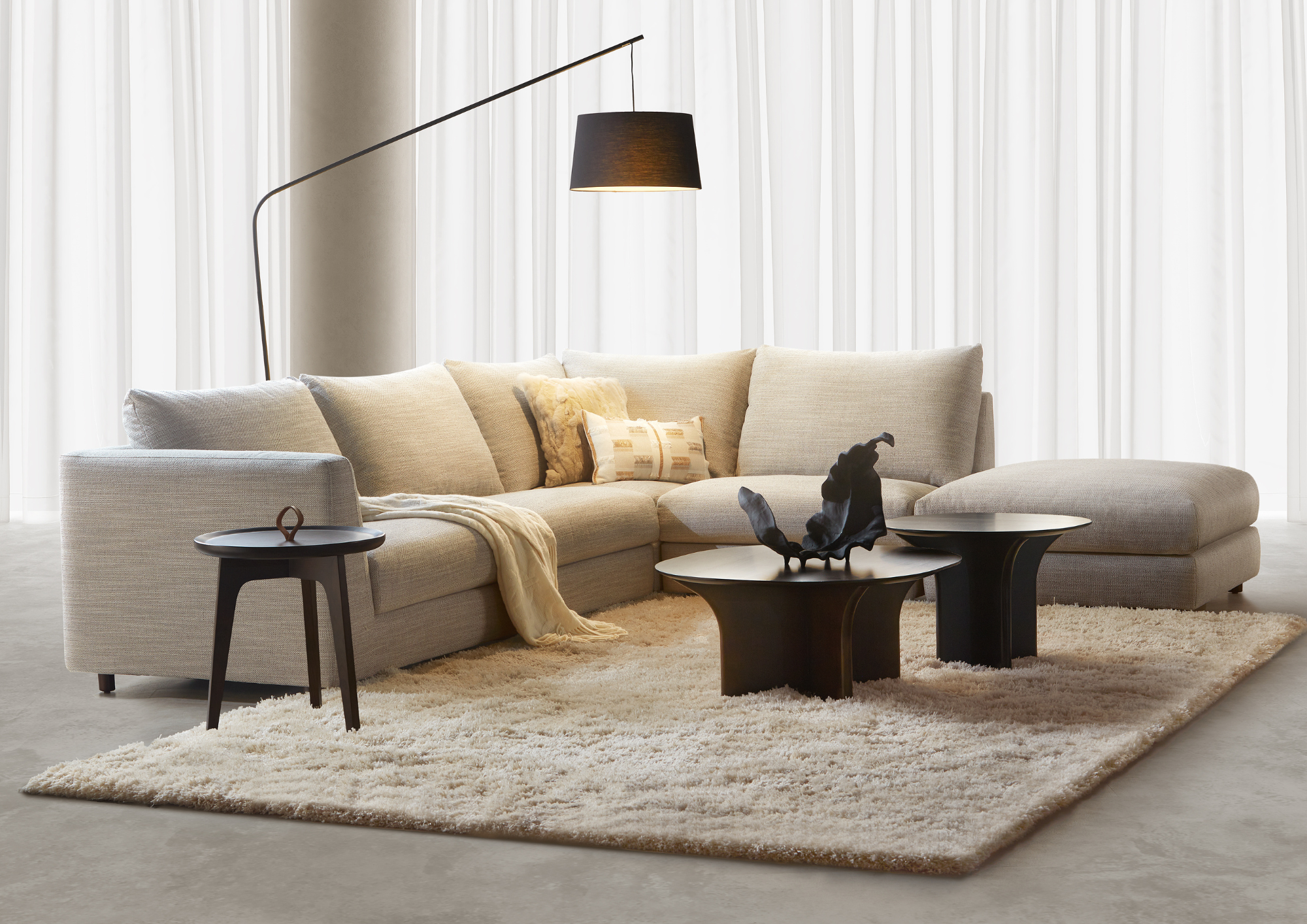 Living room with a beige L-shaped sofa, black coffee tables, a black floor lamp, cream shaggy rug, white curtains, and decorative pillows.