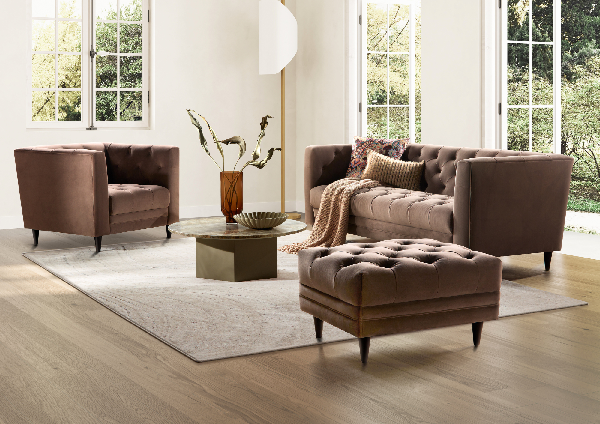 Living room with light wood flooring, a large white rug, and two armchairs. A round coffee table in the center holds a decorative bowl and a vase with artistic branches. The background features three large windows showing greenery outside, and a mode