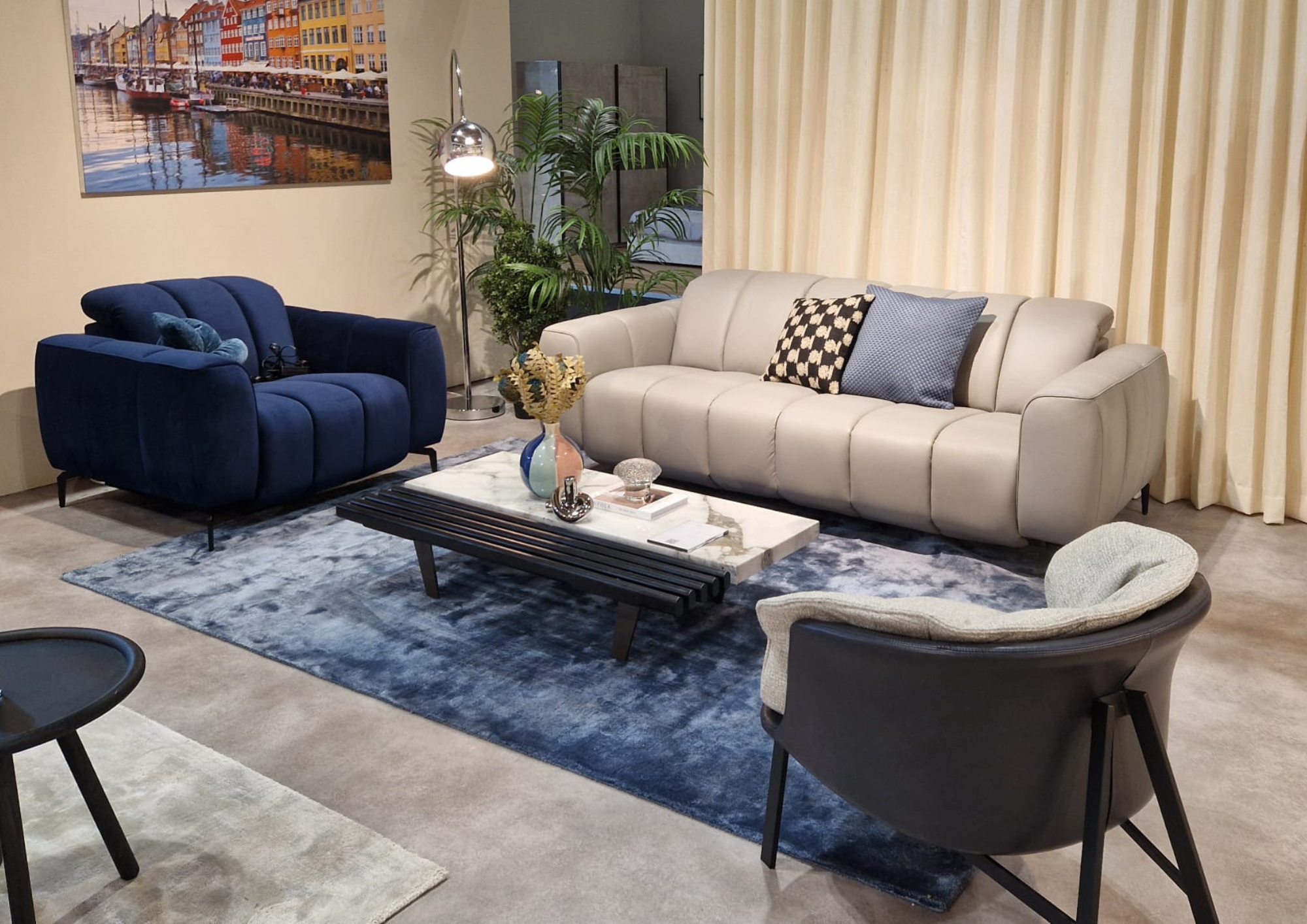 A modern living room with light beige curtains, a white sofa with patterned pillows, a navy blue armchair, a small gray armchair, a black coffee table with decorative items, a large wall art of colorful harbor buildings, a tall plant with greenery, a