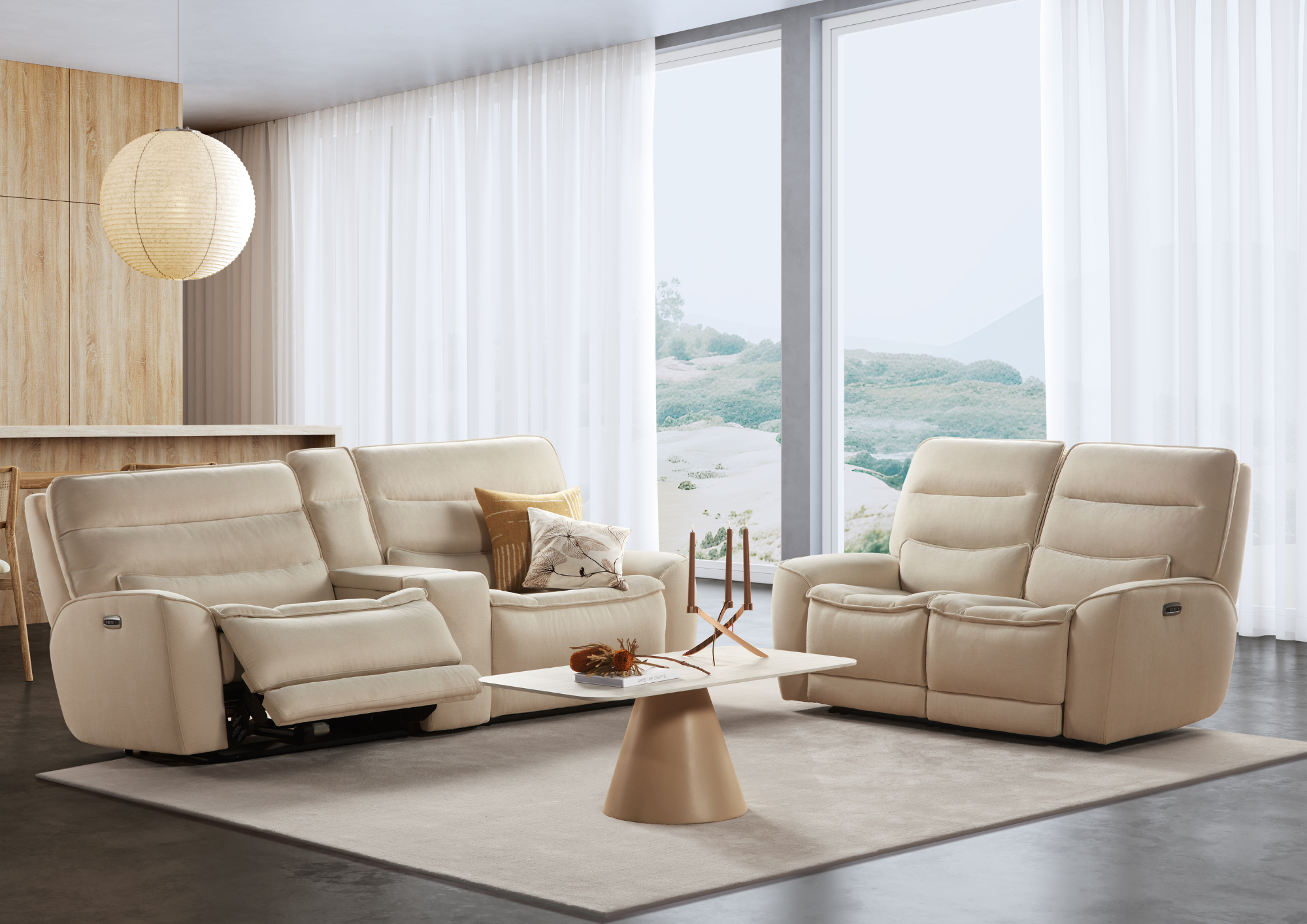 Modern living room with beige couches, beige table, large window with white curtains, wooden wall panel, white ceiling with hanging paper lantern, and outdoor view of hills.