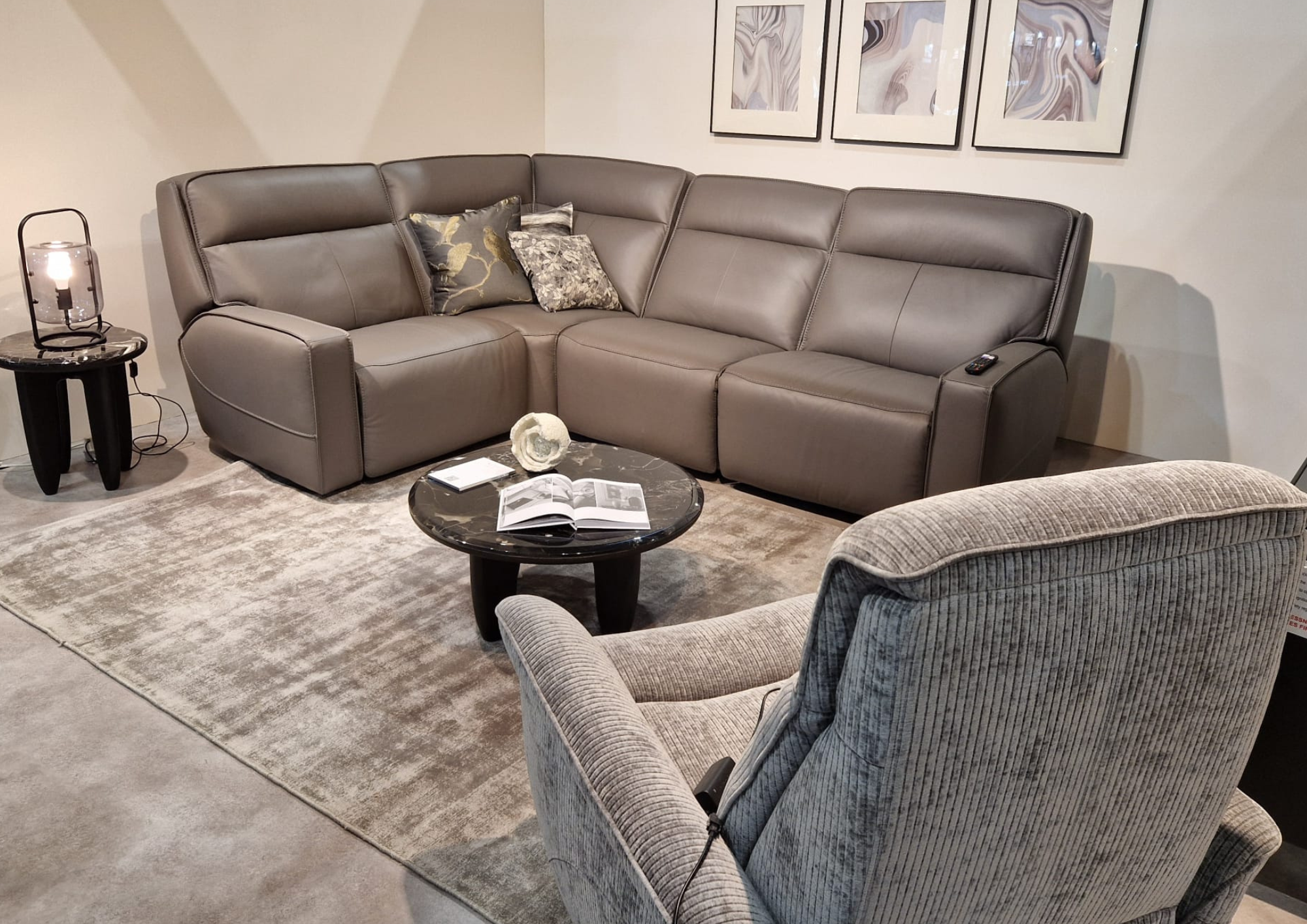 Living room with a gray leather sectional sofa, a round black marble coffee table with an open book, a gray armchair with striped upholstery, a black side table with a lantern-style lamp, and wall art with abstract patterns.