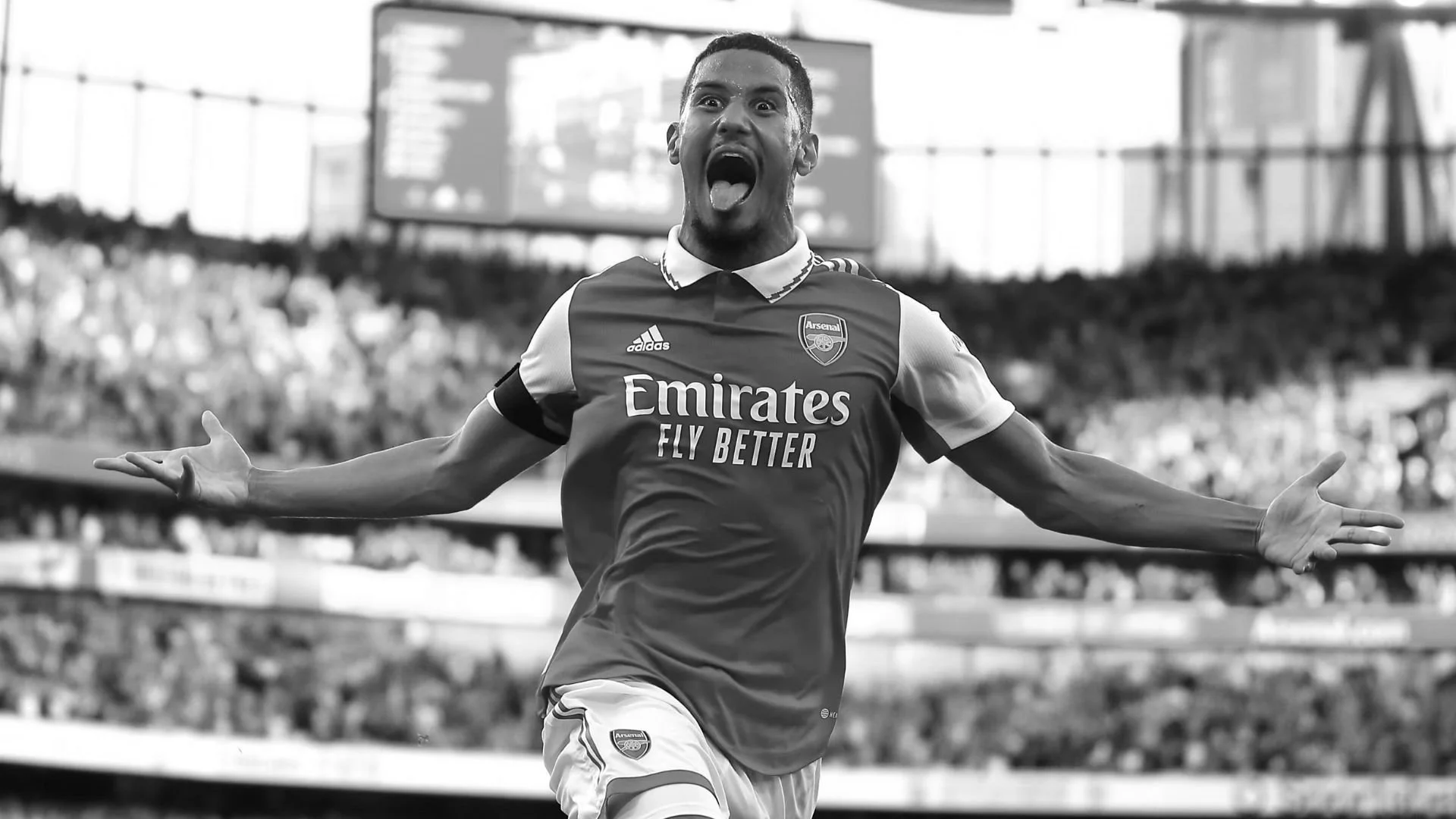 A soccer player in an Arsenal jersey celebrating on the field with arms outstretched and tongue sticking out.