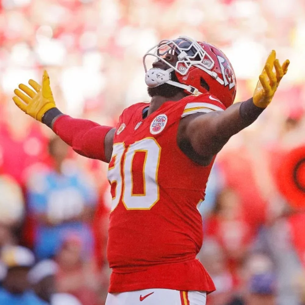 A football player wearing a red helmet, red jersey with the number 10, and yellow gloves celebrating with arms outstretched on the field.