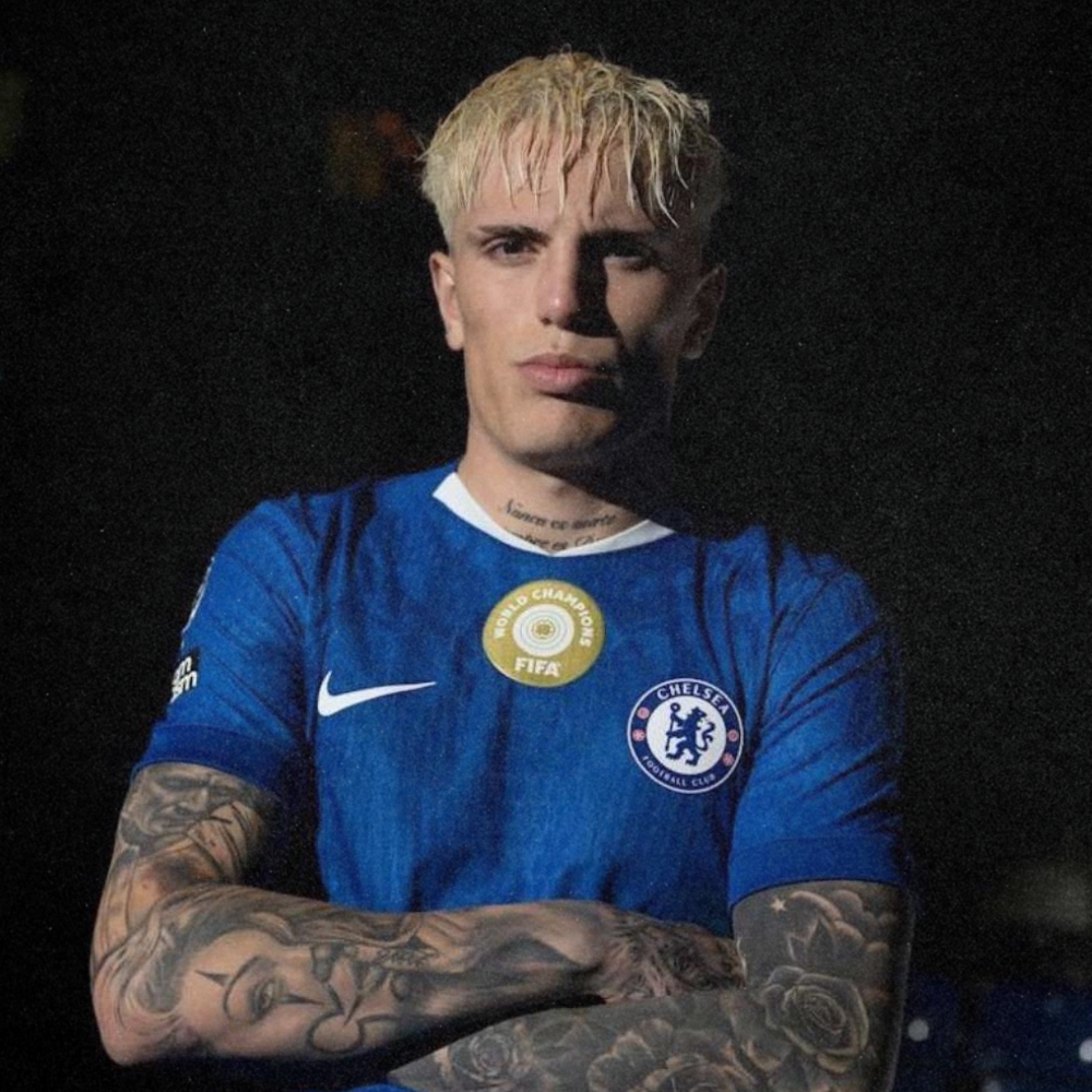 A young man with blonde hair and tattoos on his arms, wearing a blue Chelsea football jersey with FIFA and UEFA champions badges, crosses his arms and looks into the camera against a dark background.