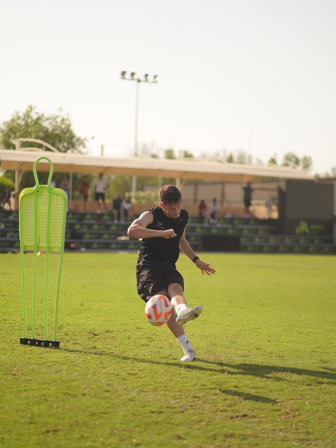 A male athlete practicing soccer outdoors, kicking the ball near training equipment on a grassy field.
