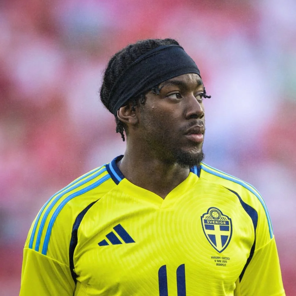 A male soccer player wearing a yellow Sweden jersey with black and blue details, a black headband, and dreadlocked hair, during a game with a blurred crowd background.