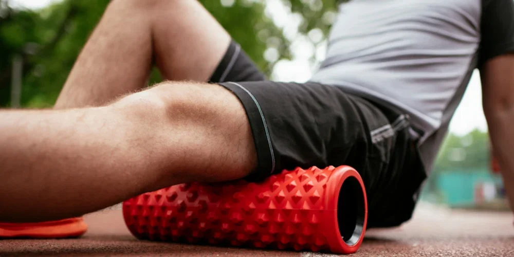 Person using a red foam roller on their thigh outdoors.