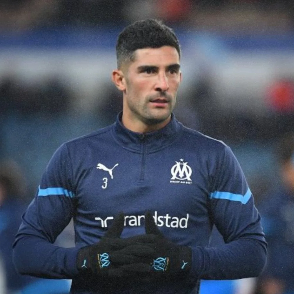A male soccer player in a navy blue uniform with the Olympique de Marseille logo, wearing gloves and standing on the field.