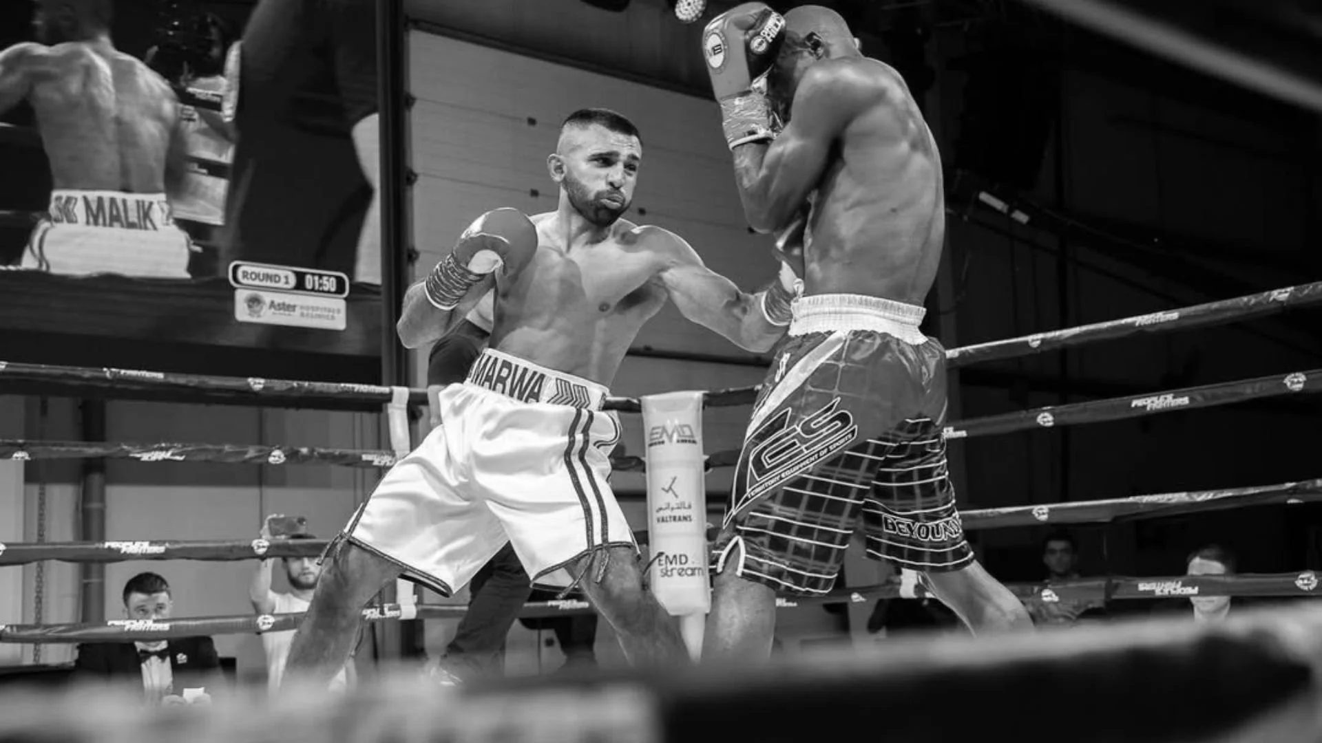 Two male boxers in a boxing ring during a match. One boxer is throwing a punch at the other, who is defending with gloves up. The image is in black and white.