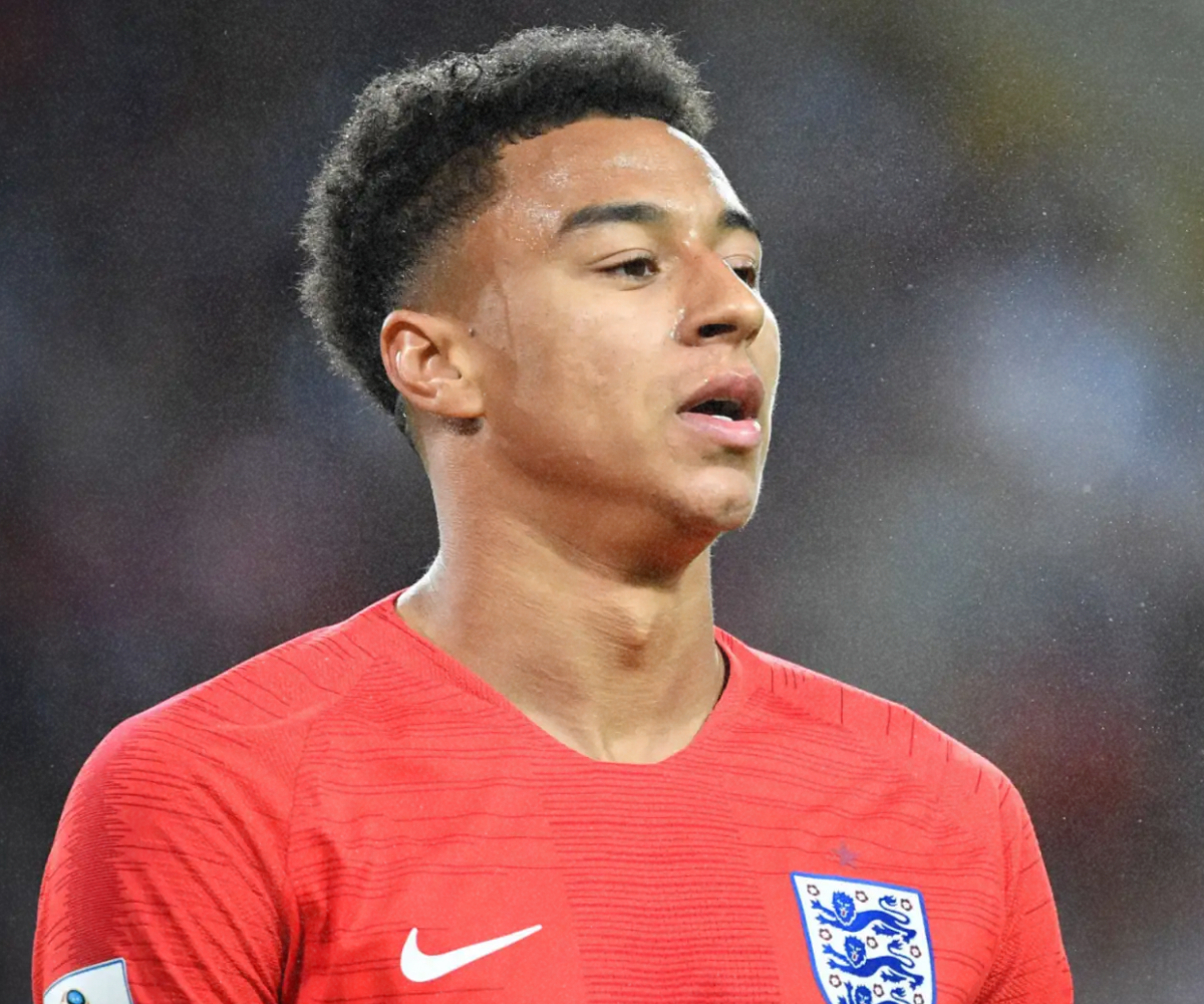 Close-up of a young male soccer player with short curly hair wearing a red England national team jersey with the Nike logo and the England crest, on a soccer field.