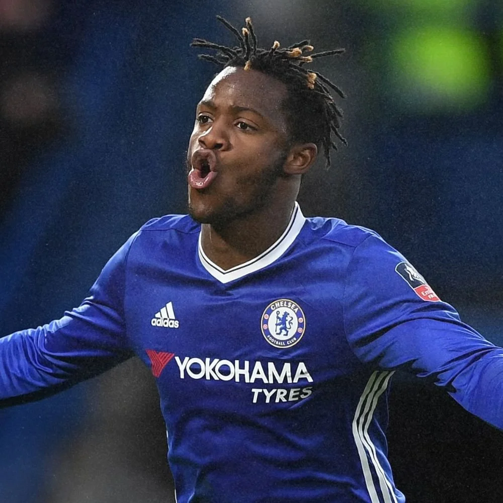 A soccer player wearing a blue Chelsea jersey with Yokohama Tyres logo, celebrating on the field with arms outstretched, puffing out his cheeks.