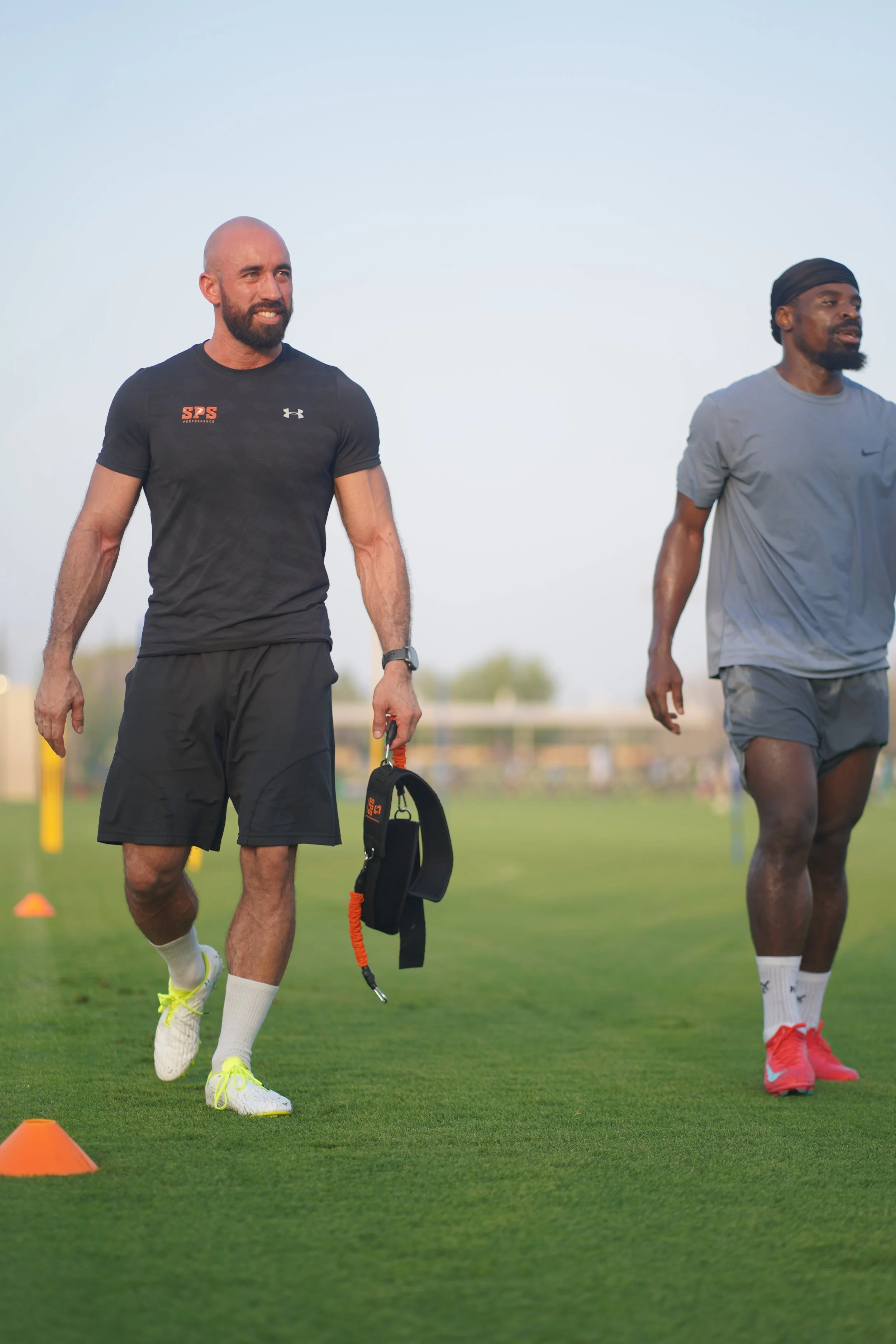 Two athletic men, one with a badge on his shirt, walking on a grassy field with orange cones, possibly during a training session.