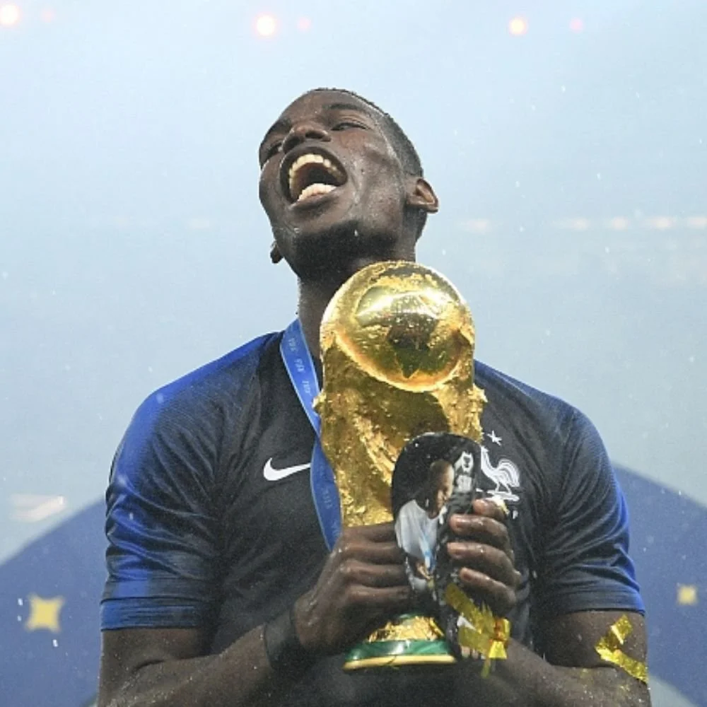 A male soccer player in a blue jersey holding a gold World Cup trophy, celebrating with an open mouth and closed eyes.
