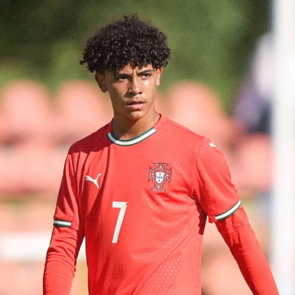 A young male soccer player with curly hair wearing a red Portugal national team jersey with the number 7, on a soccer field during a match.