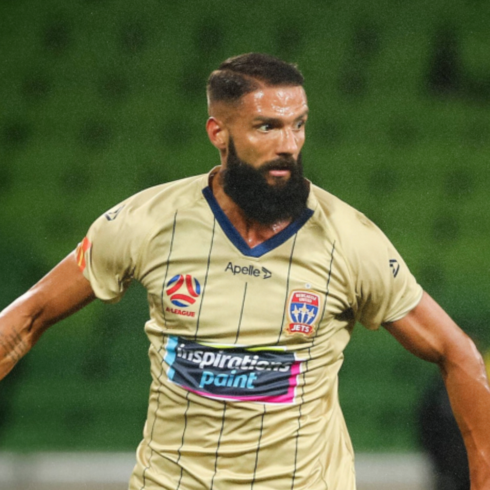 A soccer player in a beige jersey with black vertical stripes, sporting a beard and short hairstyle, on a green field.