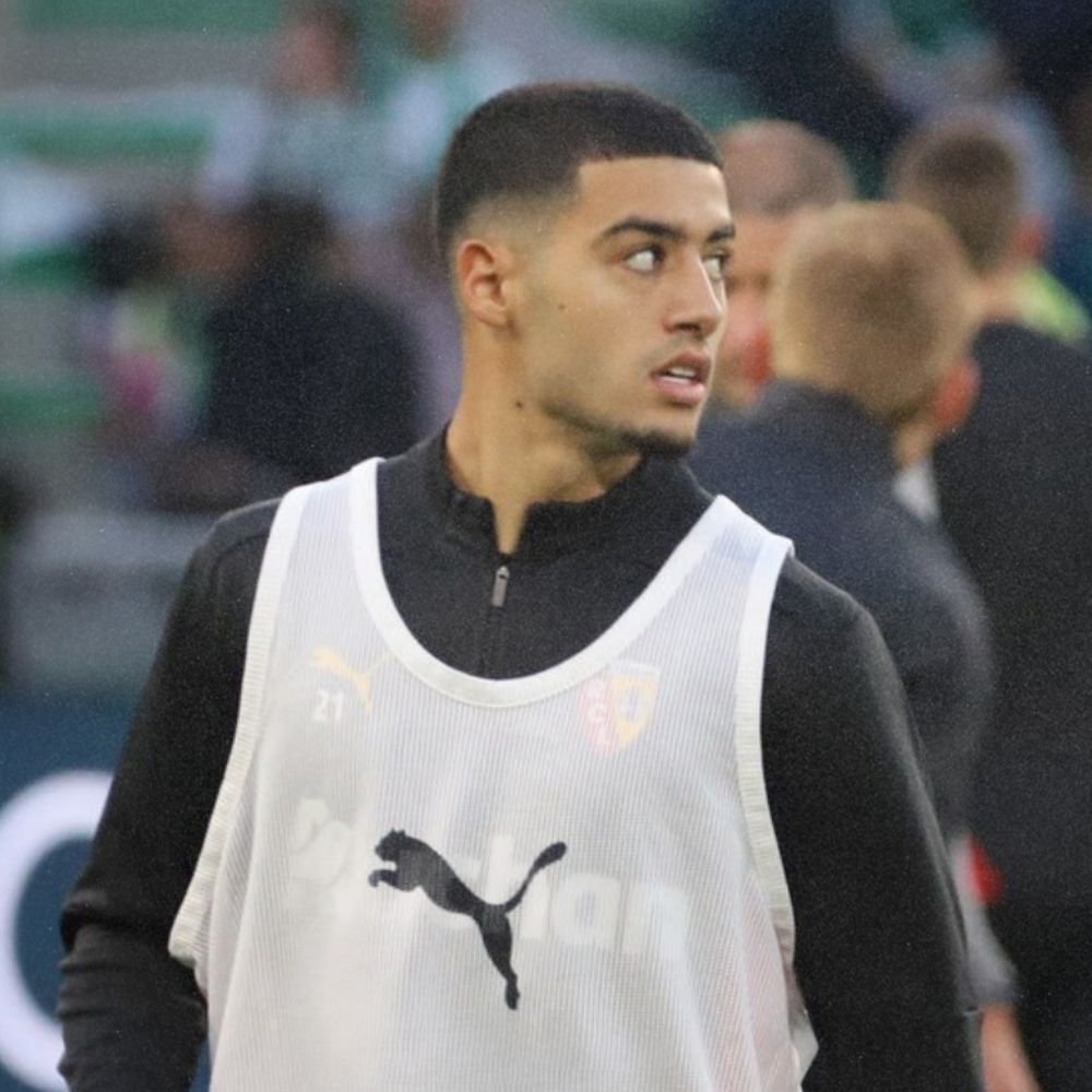 A young male soccer player with short dark hair and a beard wearing a white Puma training vest on a soccer field.