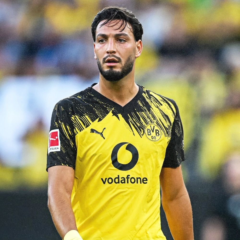 A male soccer player wearing a black and yellow Borussia Dortmund jersey with the Vodafone logo, standing on a soccer field with a blurred background of spectators.