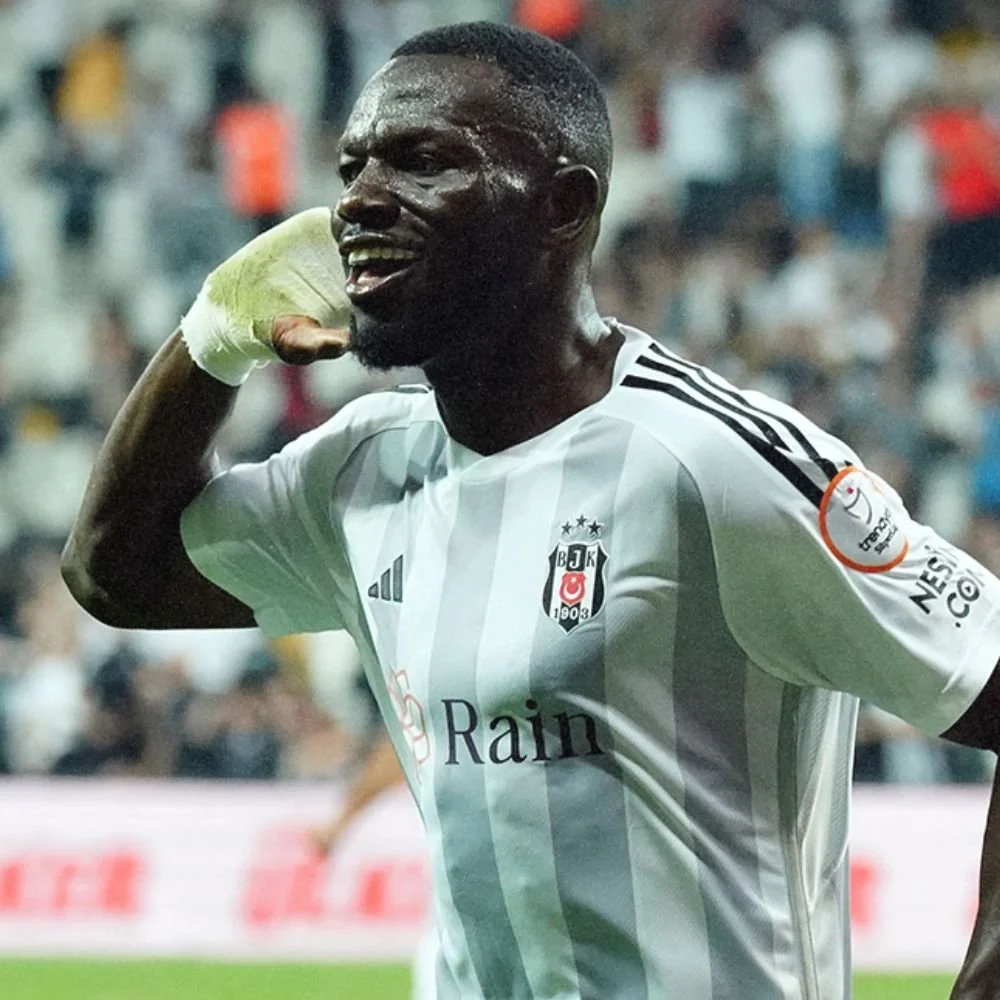 A soccer player celebrating after scoring a goal, wearing a white jersey with black stripes and logos, and a yellowish-green glove covering his left hand.