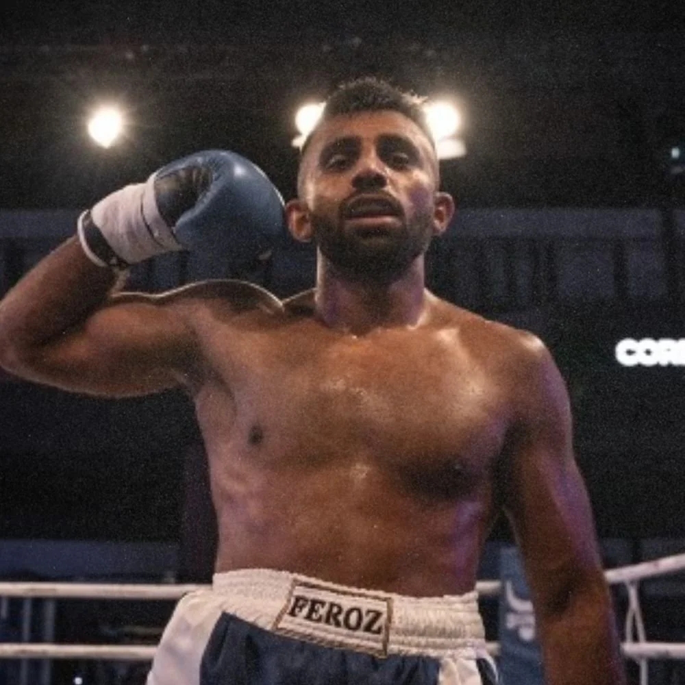 A shirtless male boxer with a beard and short hair, wearing boxing trunks and gloves, stands in a boxing ring with his hand on the back of his head, looking contemplative.