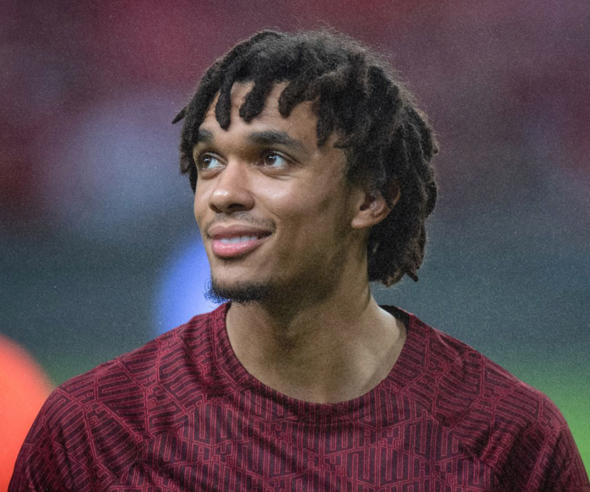 A young man with brown skin and dreadlocks wearing a red shirt, smiling and looking slightly to the side.
