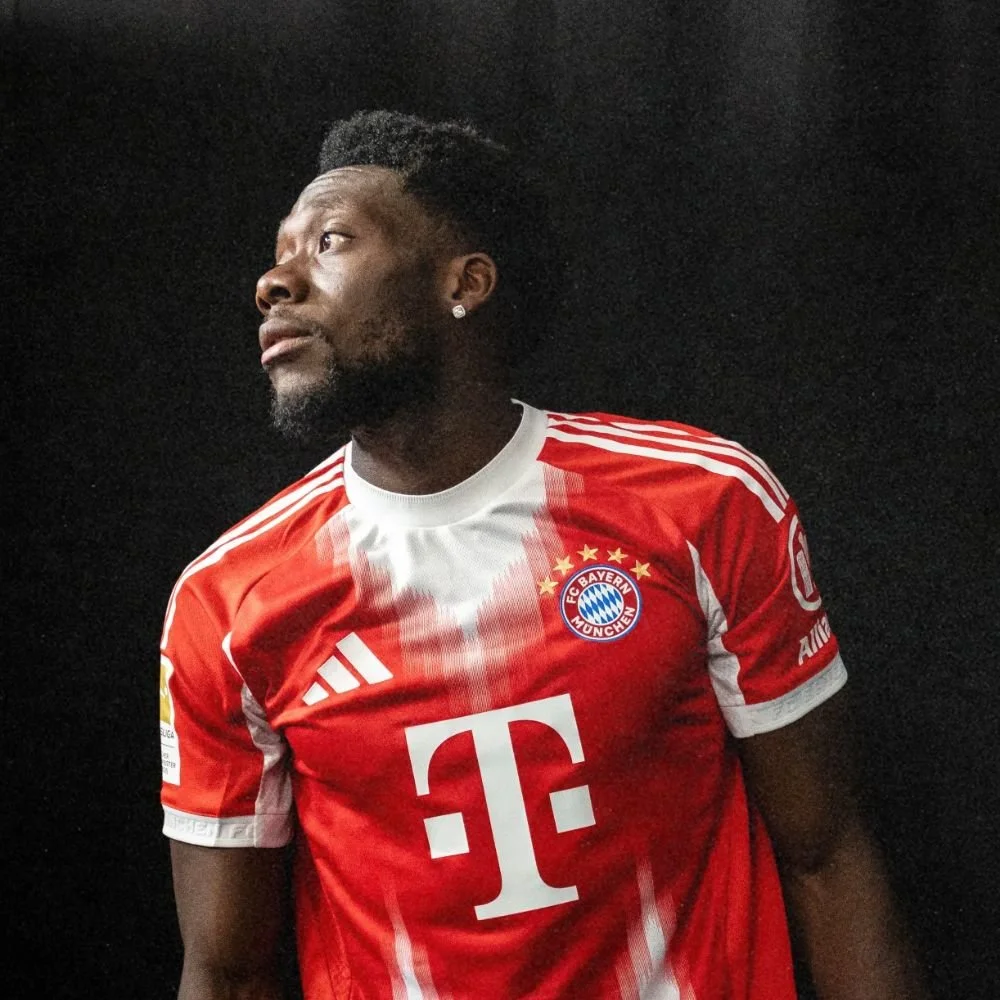 A man wearing a red Bayern Munich soccer jersey with white accents, standing against a dark background.