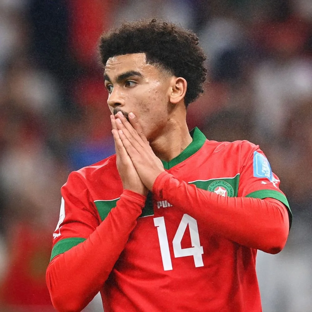 A young male soccer player wearing a red jersey with the number 14 on it, holding his hands together in front of his mouth in a gesture of concern or prayer during a game.