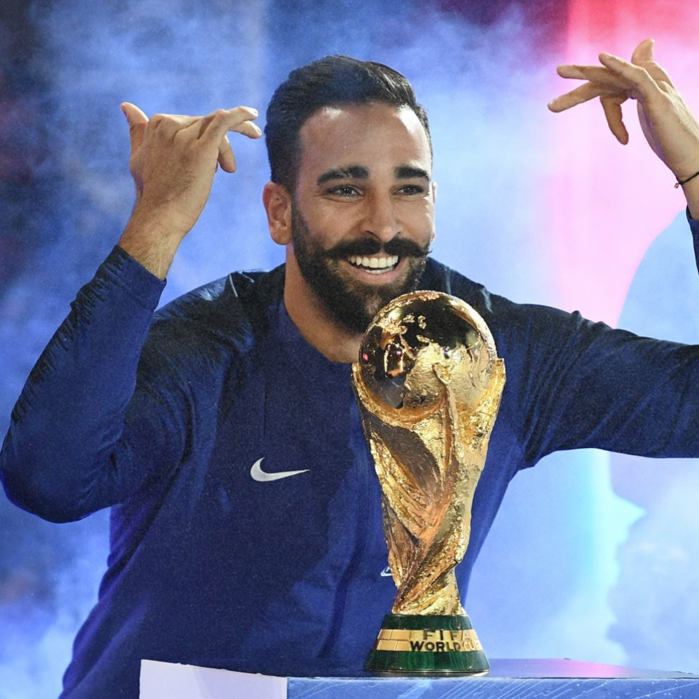 A man with dark hair and a beard is smiling and posing with a golden FIFA World Cup trophy, wearing a navy blue athletic jacket.