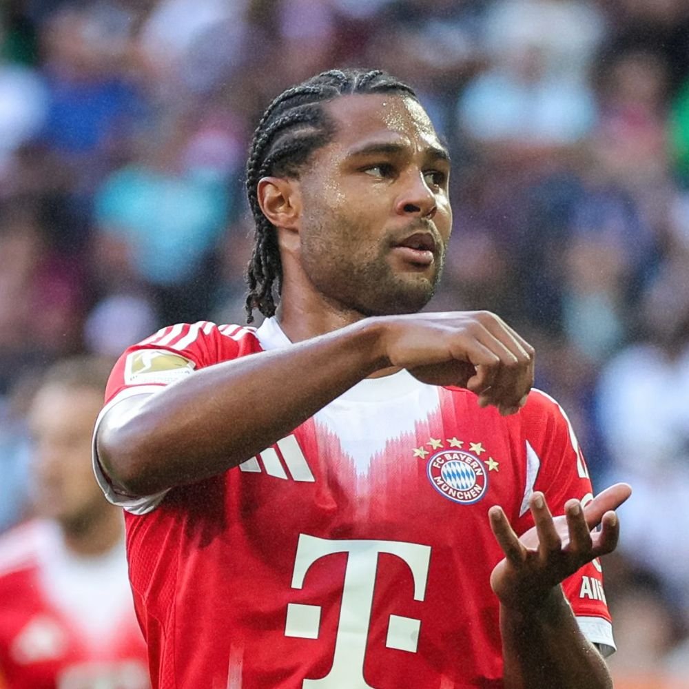 A soccer player in a red Bayern Munich jersey is performing a celebratory gesture on the field during a match.