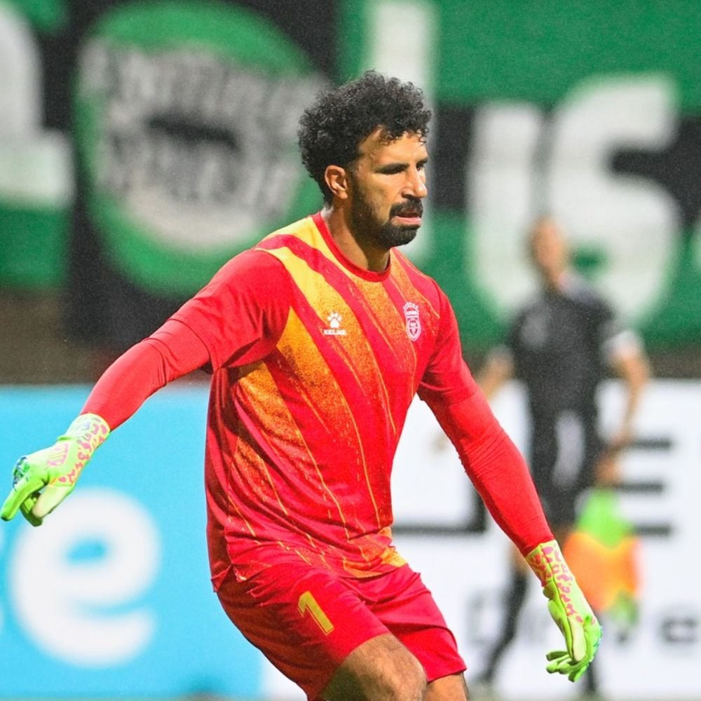 Soccer player in red and yellow uniform with gloves, standing on the field with a serious expression.