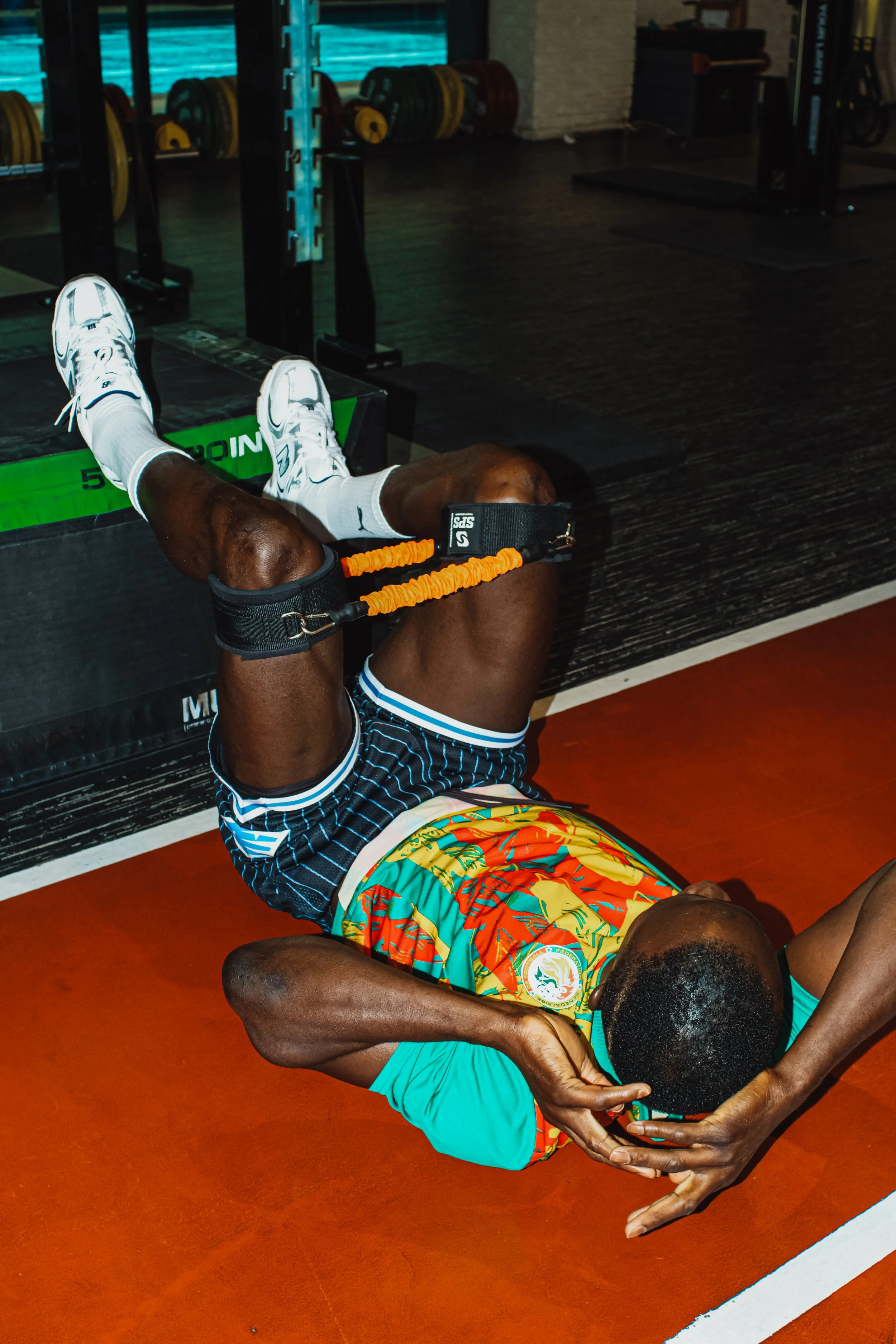 A person lying on the gym floor, wearing gloves, colorful athletic clothing, and white sneakers, with their knees raised and secured with resistance bands.