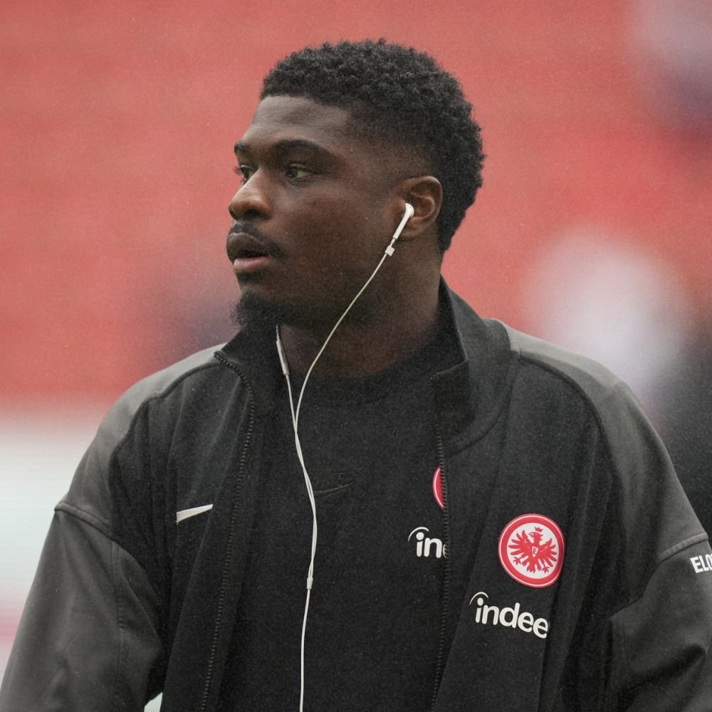 A young man with short curly hair and a dark complexion is wearing a black jacket and black shirt with logos, and white earphones, outdoors with a blurred red background.