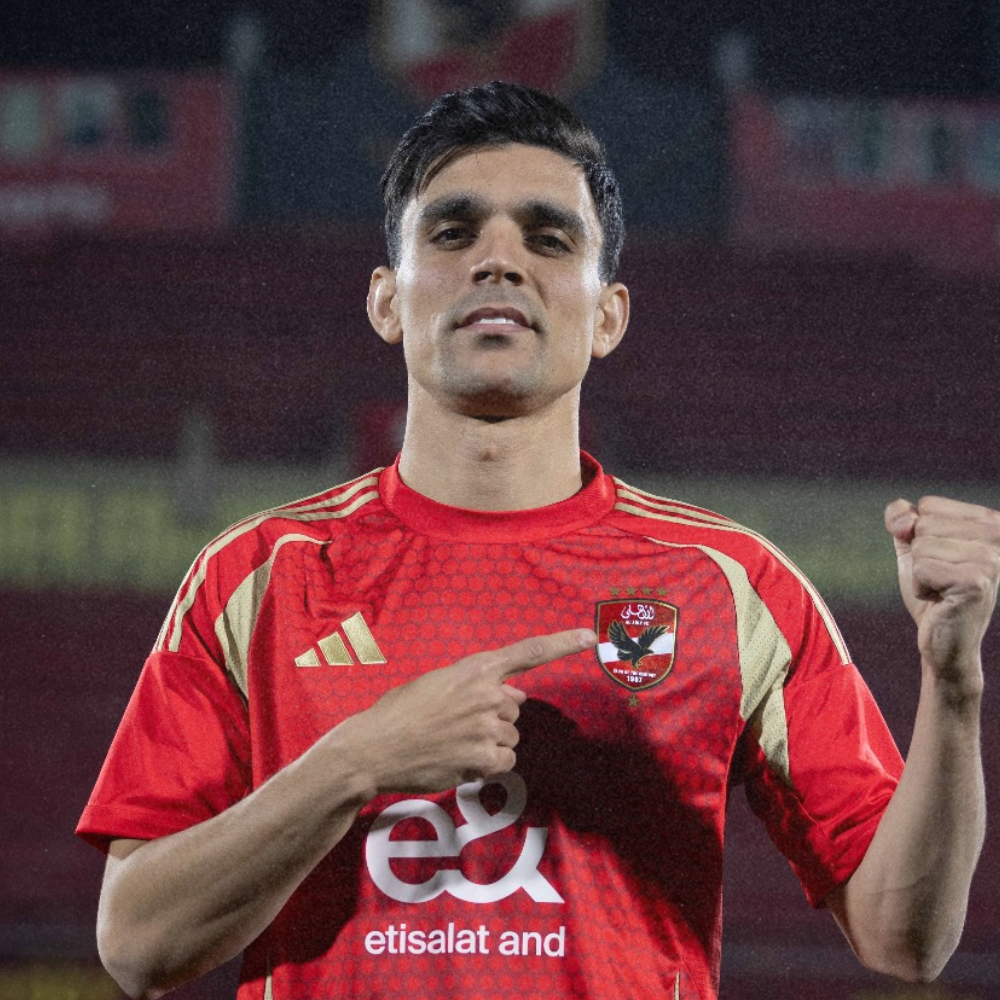Soccer player in red and gold jersey pointing to a crest on his chest with his right hand and making a fist with his left hand.