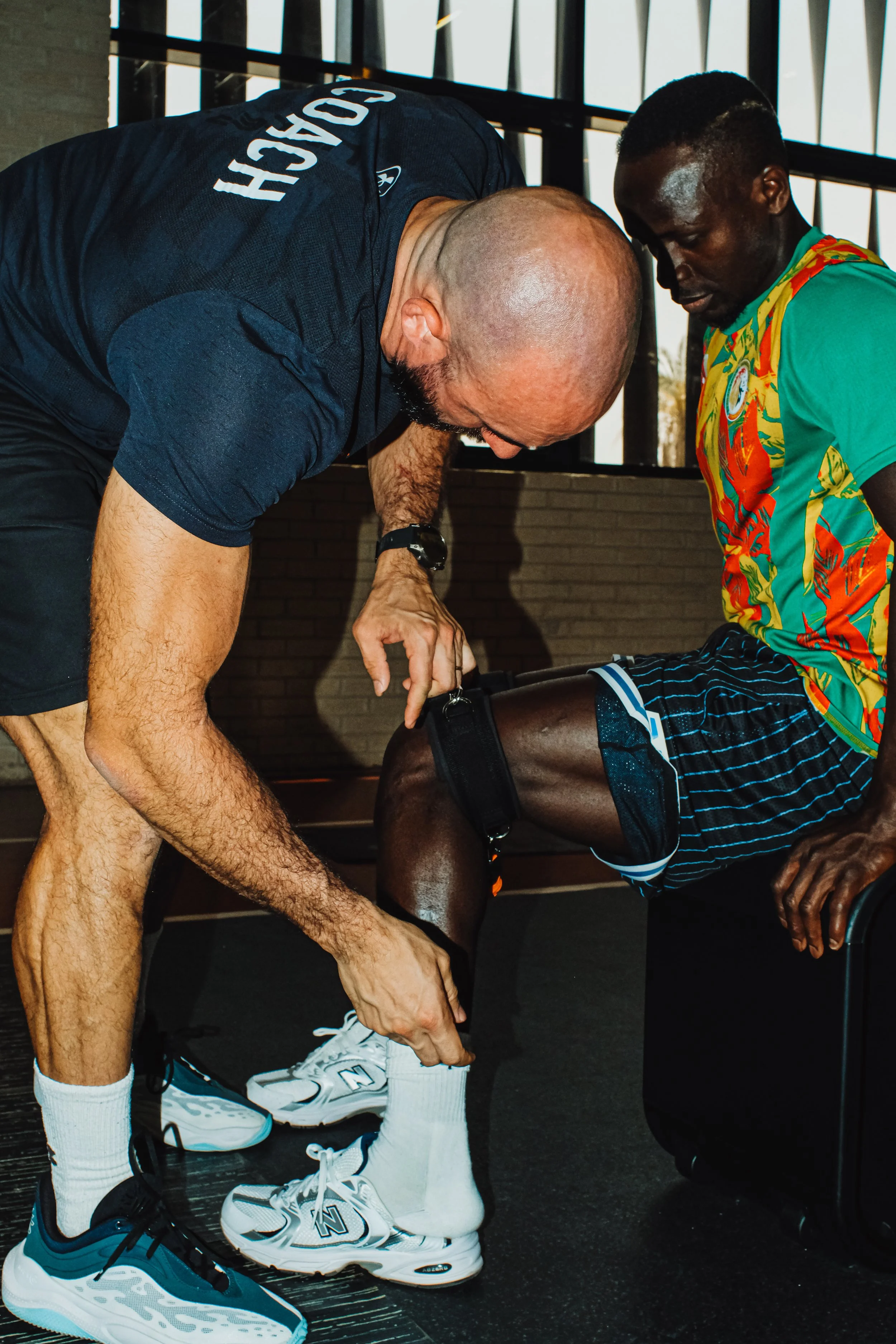 A man helps a runner put on his sock before a workout or race in an indoor setting.
