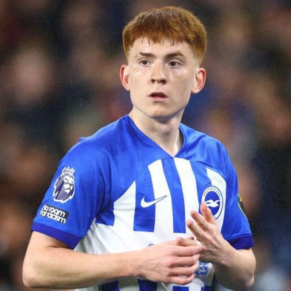 A young male soccer player with red hair wearing a blue and white striped jersey with a lion crest, Nike logo, and Premier League patch on the sleeve, standing on a soccer field during a match.