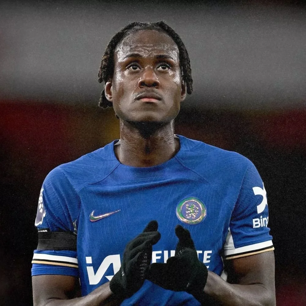 A male soccer player in a blue jersey with the Chelsea Football Club logo, standing on the field with hands clasped in front, looking upward.