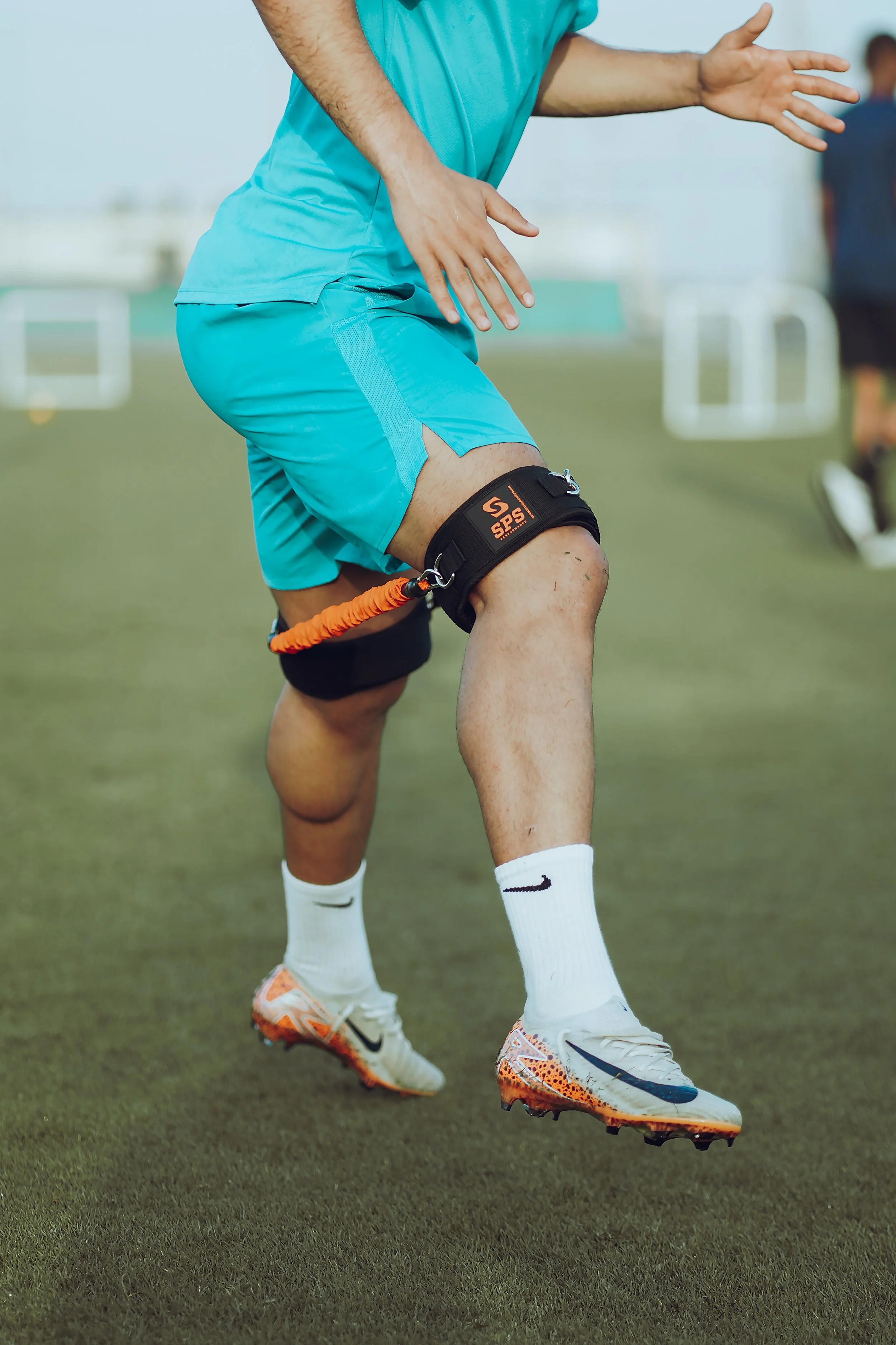 An athlete wearing teal sportswear using a resistance band around his thigh during training on a grassy field.