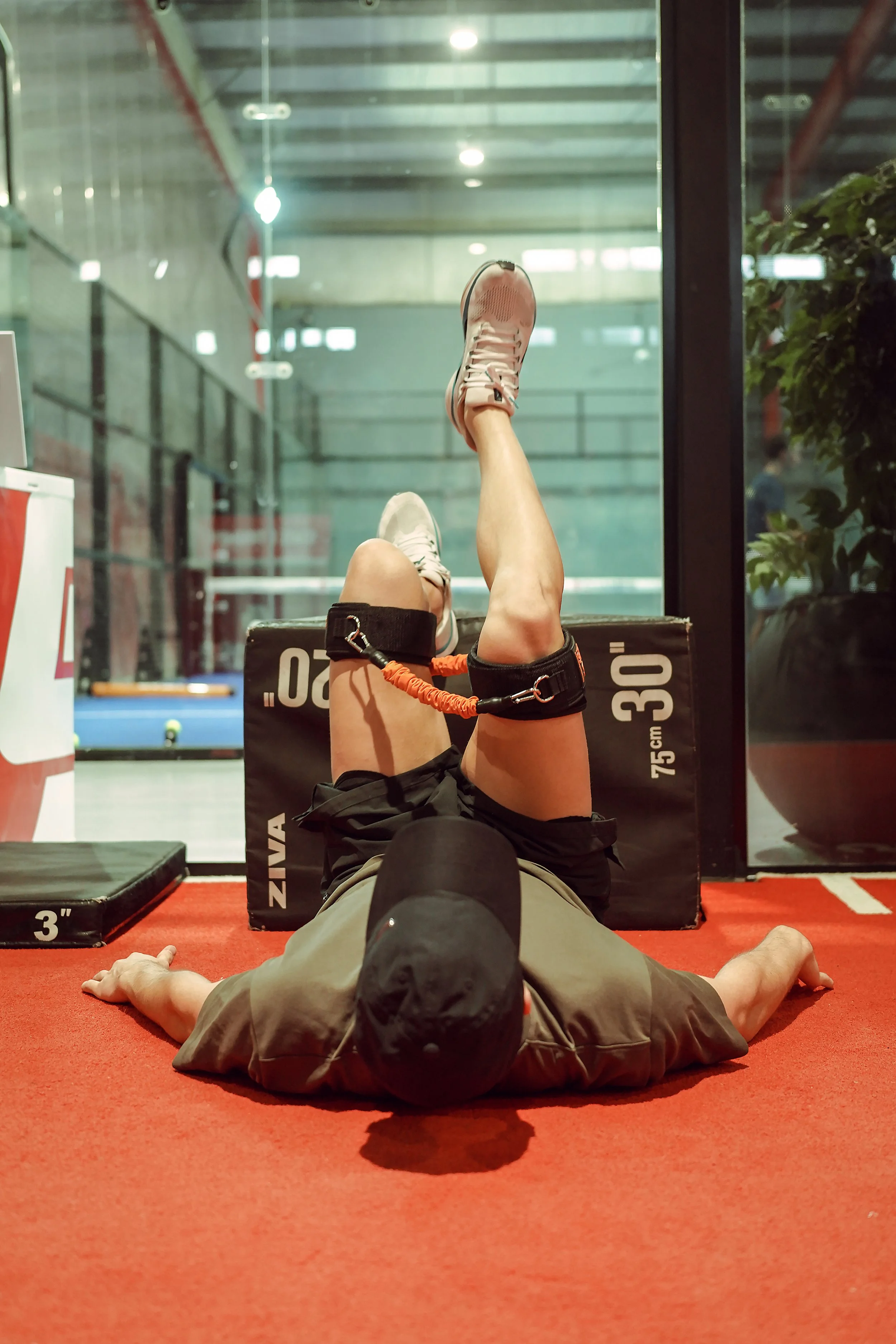 A man lying on the gym floor with an ankle strap resistance band around his right ankle, performing leg exercises. He is wearing a black cap, gray shirt, and black shorts, with gym equipment and a glass wall in the background.