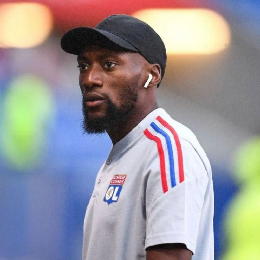 A man in a white sports jersey with red and blue stripes on the shoulders, wearing a black cap and AirPods, looking to the side against a blurred background of lights.