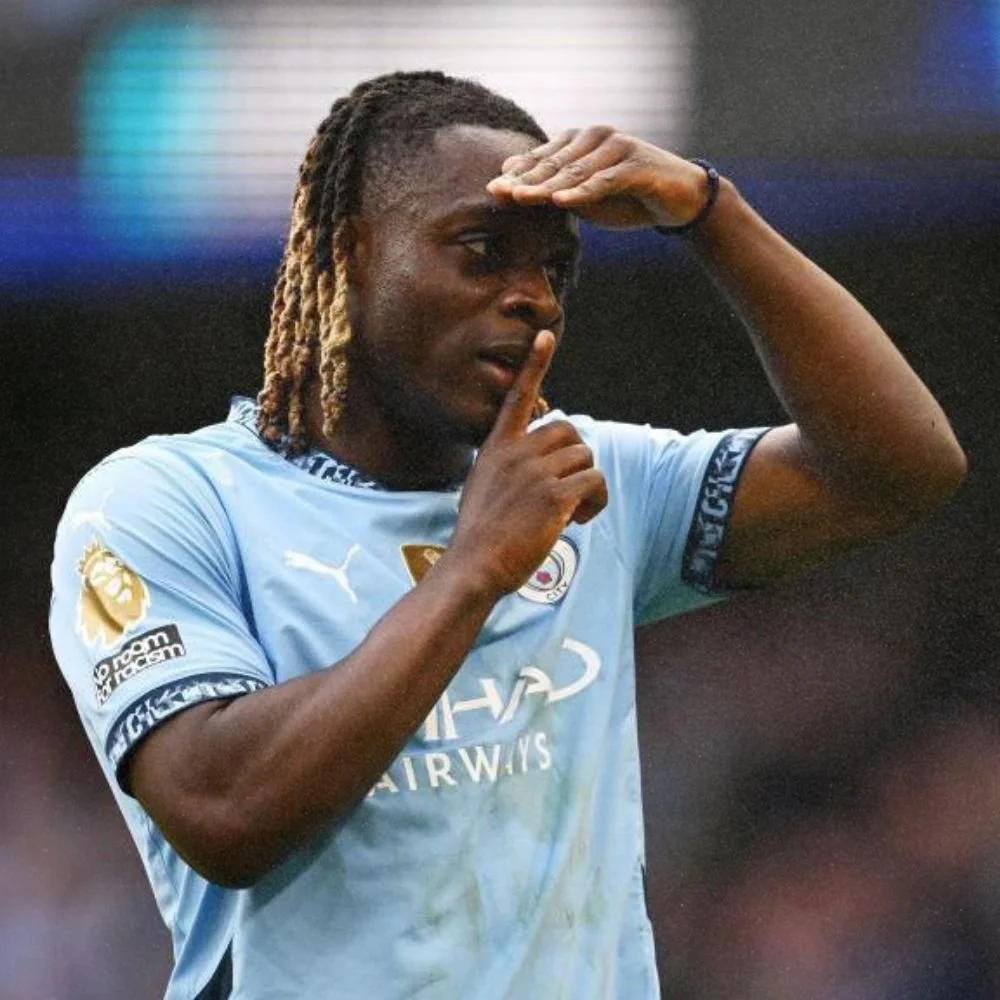 Soccer player wearing a light blue Manchester City jersey making a shushing gesture with his finger to his lips, holding his hand to his forehead.