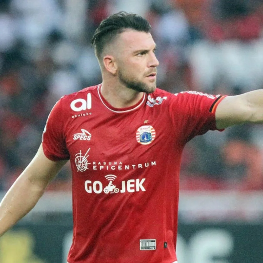 A male soccer player with a beard and styled hair wearing a red jersey with various sponsors and an emblem, on a soccer field during a game.