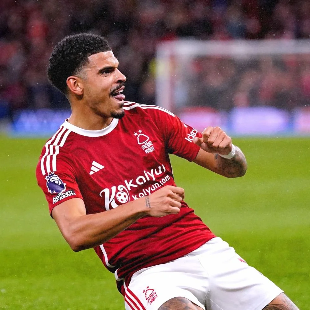 A soccer player wearing a red jersey with white stripes on the shoulders and white shorts, celebrating on the field with clenched fists and a joyful expression.