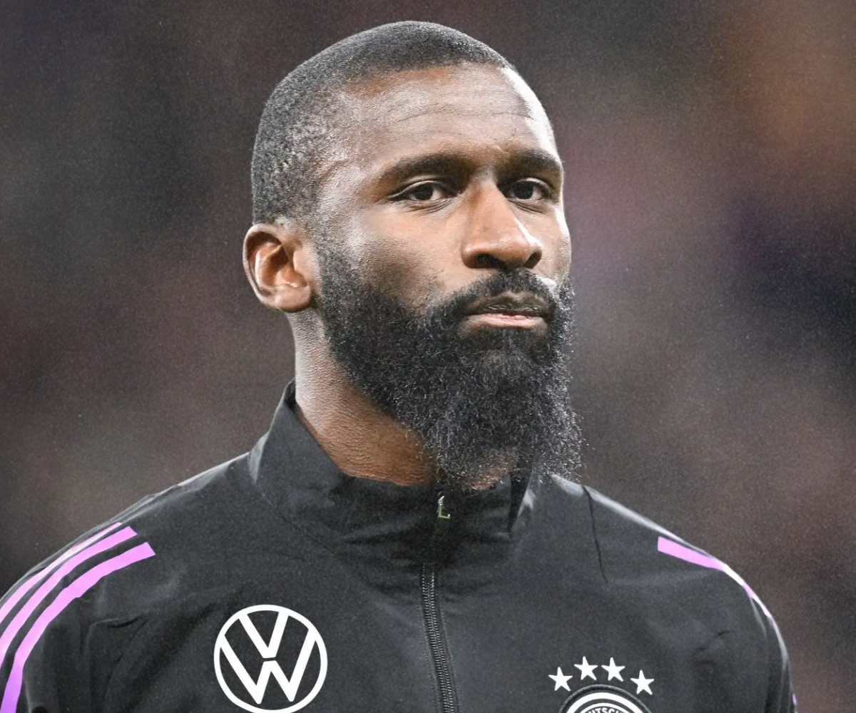 A professional soccer player with a beard wearing a black training jacket with purple stripes, featuring the Volkswagen logo and German national team emblem, looking serious.