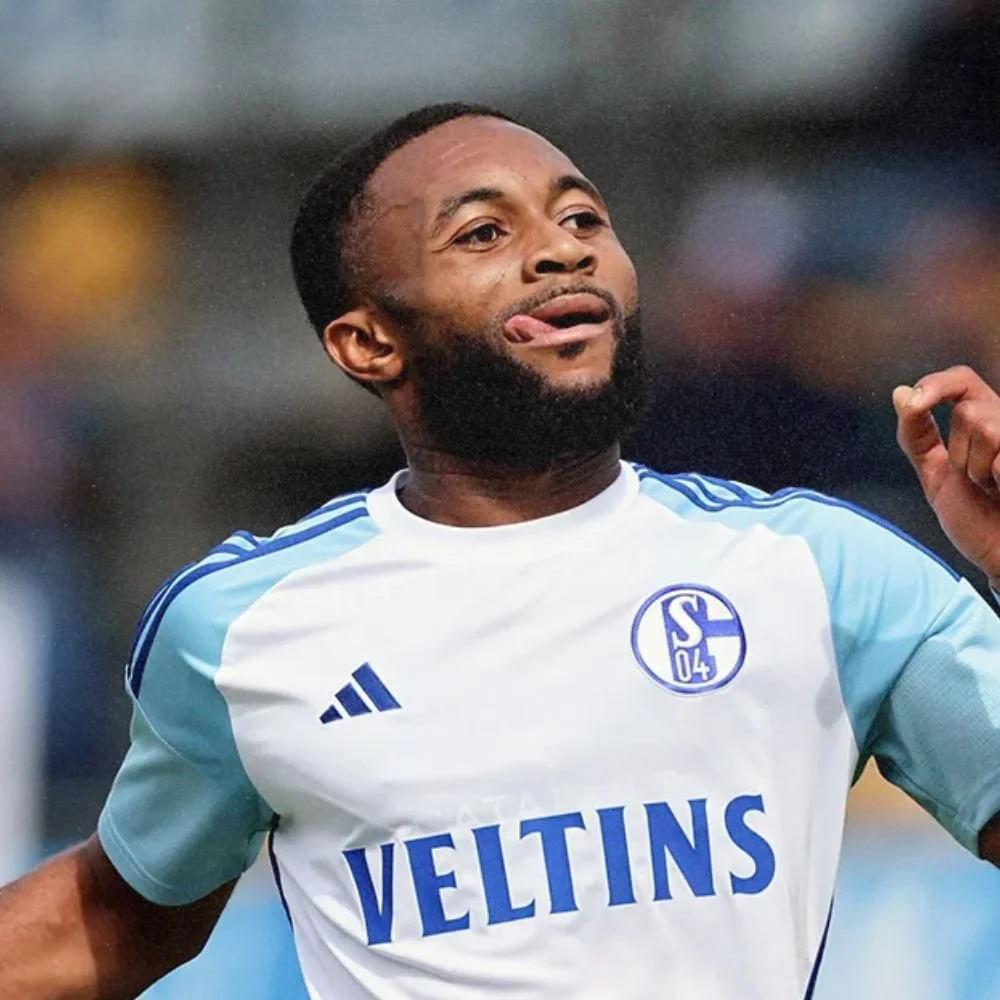 Soccer player wearing a white and light blue jersey with the logo 'S04' and the word 'VELTINS,' celebrating during a game.