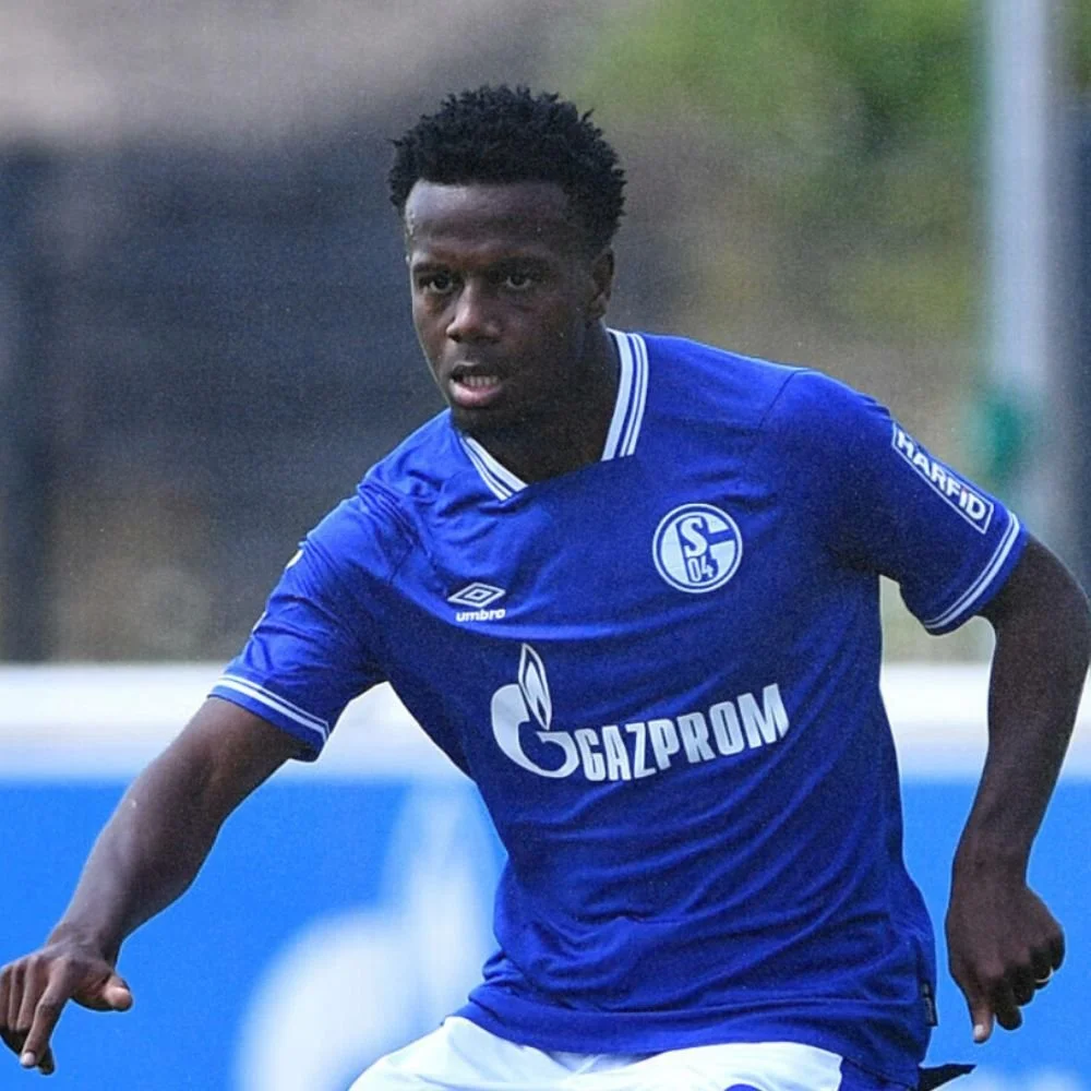 A soccer player wearing a blue jersey with the Schalke 04 logo and Gazprom sponsor, running on the field during a game.